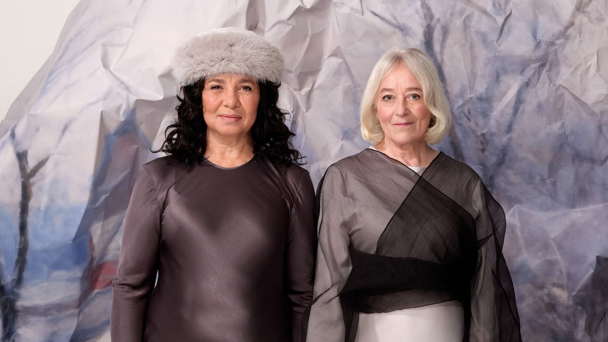 Twee vrouwen staan naast elkaar met hun vingers in elkaar verweven. Achter hen staat een ijsberg van papier. De linker vrouw heeft donkerbruin krullend haar, draagt een grijze muts en een lange bruine jurk. De rechter vrouw heeft kort, stijl en blond haar en draagt een lange zwart/witte jurk.