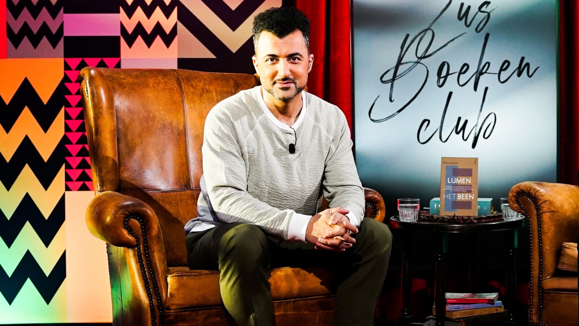 Man met zwart haar en een baardje zit met zijn handen gevouwen op een stoel voor een kleurijk decor