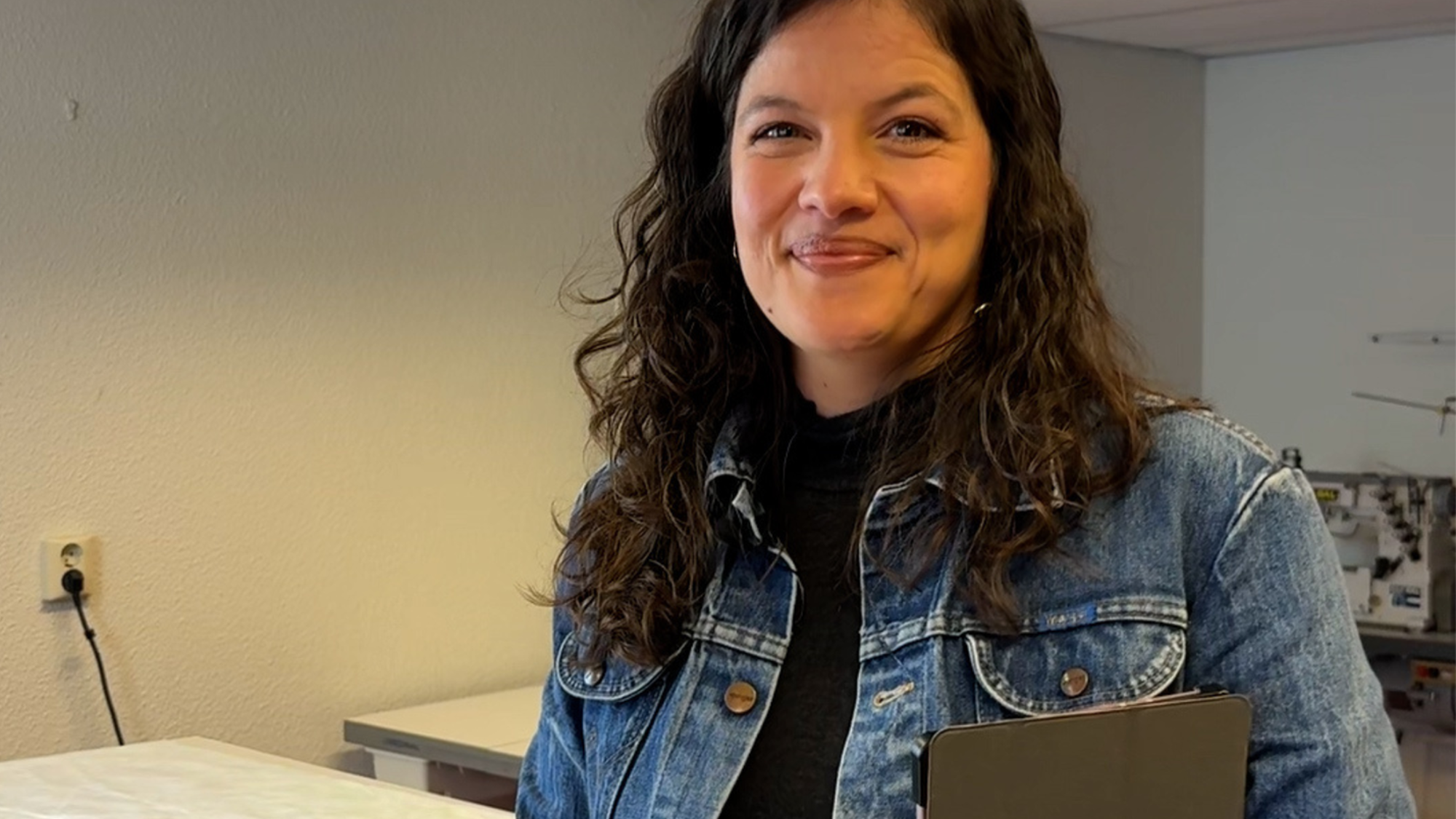 Foto van Lorena in haar atelier. Ze heeft bruin haar loshangend en een spijkerpak aan. Ze heeft een tablet en pen in haar hand en lacht naar de camera.