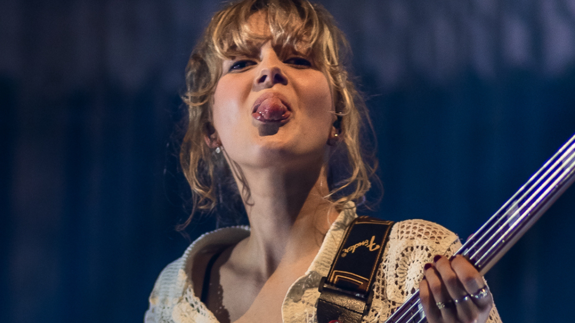 Een foto van Pieteke, een blonde meid met pony. Ze staat op een podium met een basgitaar in haar handen en steekt haar tong uit.