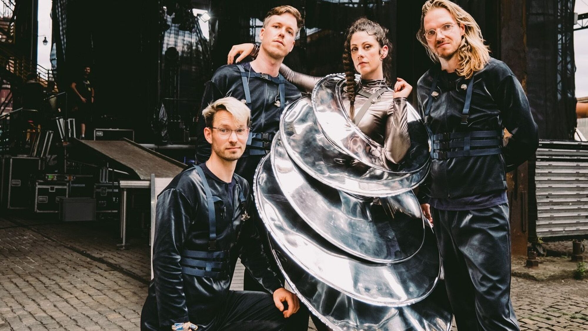 Een foto van vier mensen: links twee witte mannen, in het midden een witte vrouw en rechts ook een witte man. De mannen links hebben beide kort haar, dragen een blauw harnas en een zwart pak. De man rechts draagt dezelfde outfit en heeft lang blond haar. De vrouw in het midden draagt een outfit die lijkt te bestaan uit verschillende zilveren schotels. Haar lange bruine haar draagt ze in een vlecht. Hun blikken zijn serieus.