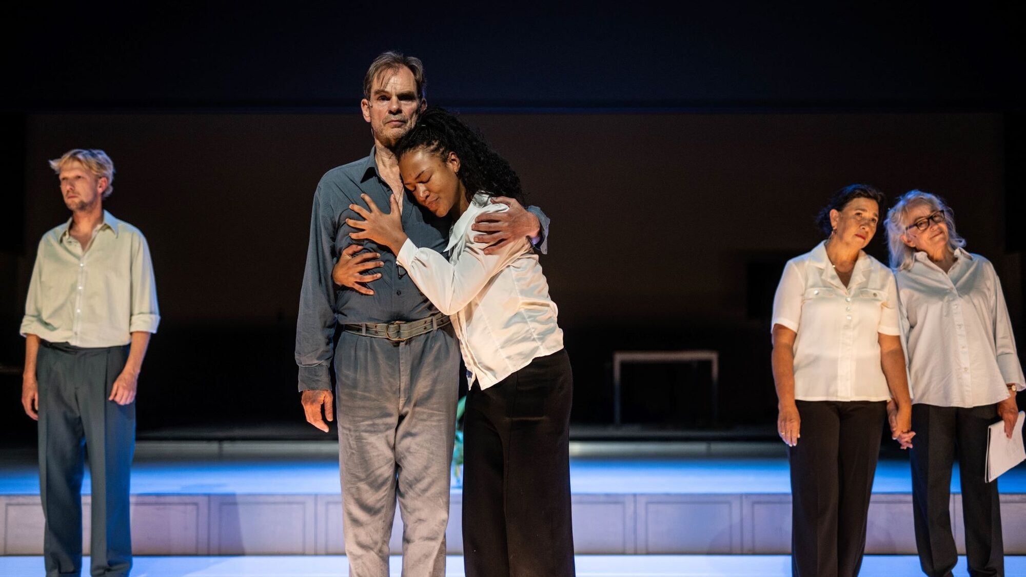 Vijf mensen staan op een podium. Links staat een blonde manier met een overhemd en een blauwe broek voor zich uit te staren. In het midden omhelzen twee mensen elkaar: links een man en rechts een vrouw. De man staart voor zich uit en de vrouw kijkt geëmotioneerd. Rechtsachter staan twee vrouwen die elkaars hand vasthouden. Ze dragen beide een witte blouse en zwarte broek. De linker vrouw kijkt peinzend voor zich uit en de rechter kijkt vrolijk.