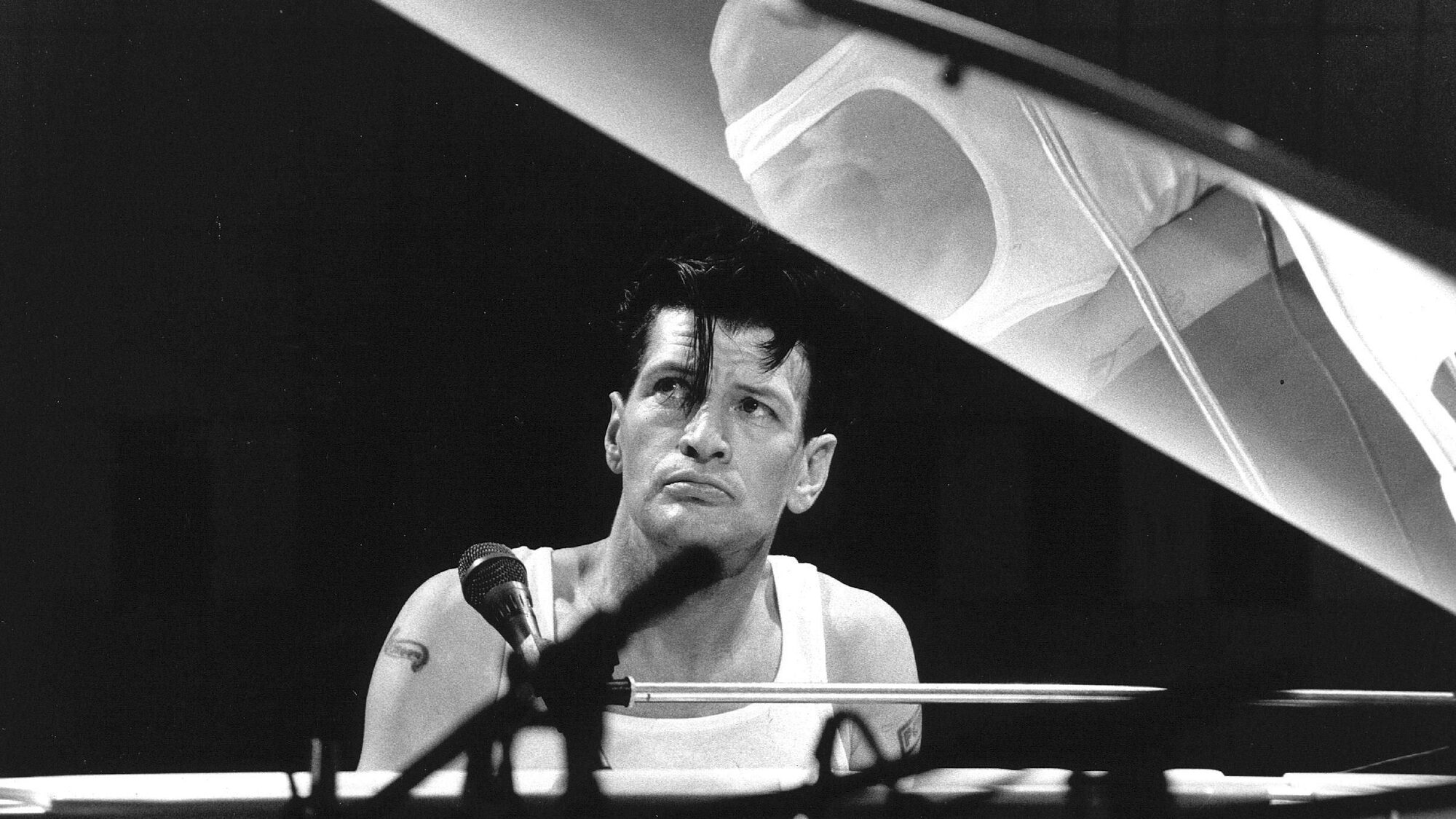 Herman Brood zit peinzend achter een vleugel in een wit hemd