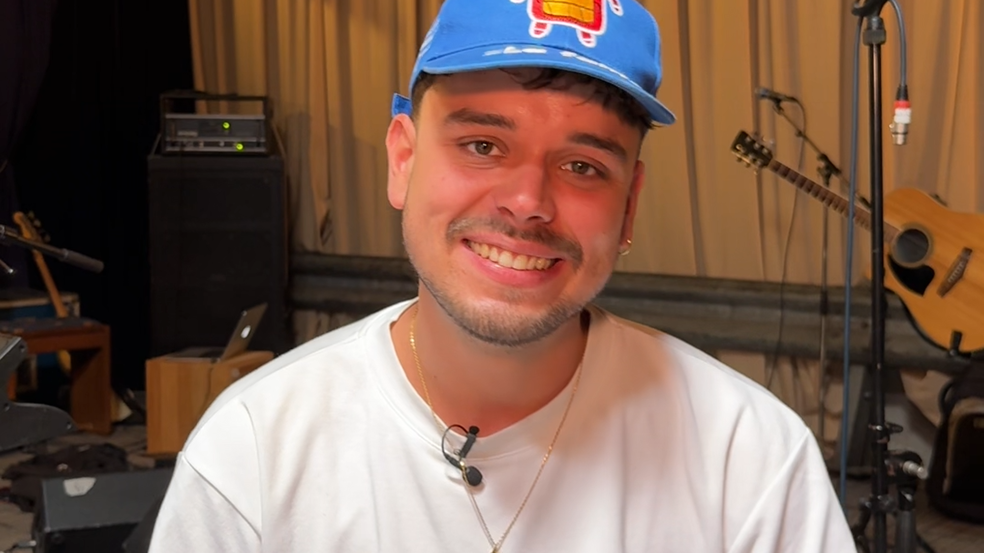 Een foto van de artiest Quique, die getipt wordt in dit stukje tekst. Het is ene jonge jongen met een wit shirt en een pet en hij kijkt lachend de camera in. Hij zit in een repetitieruimte, het is een beetje donker en er hangen gelige gordijnen voor de muur.