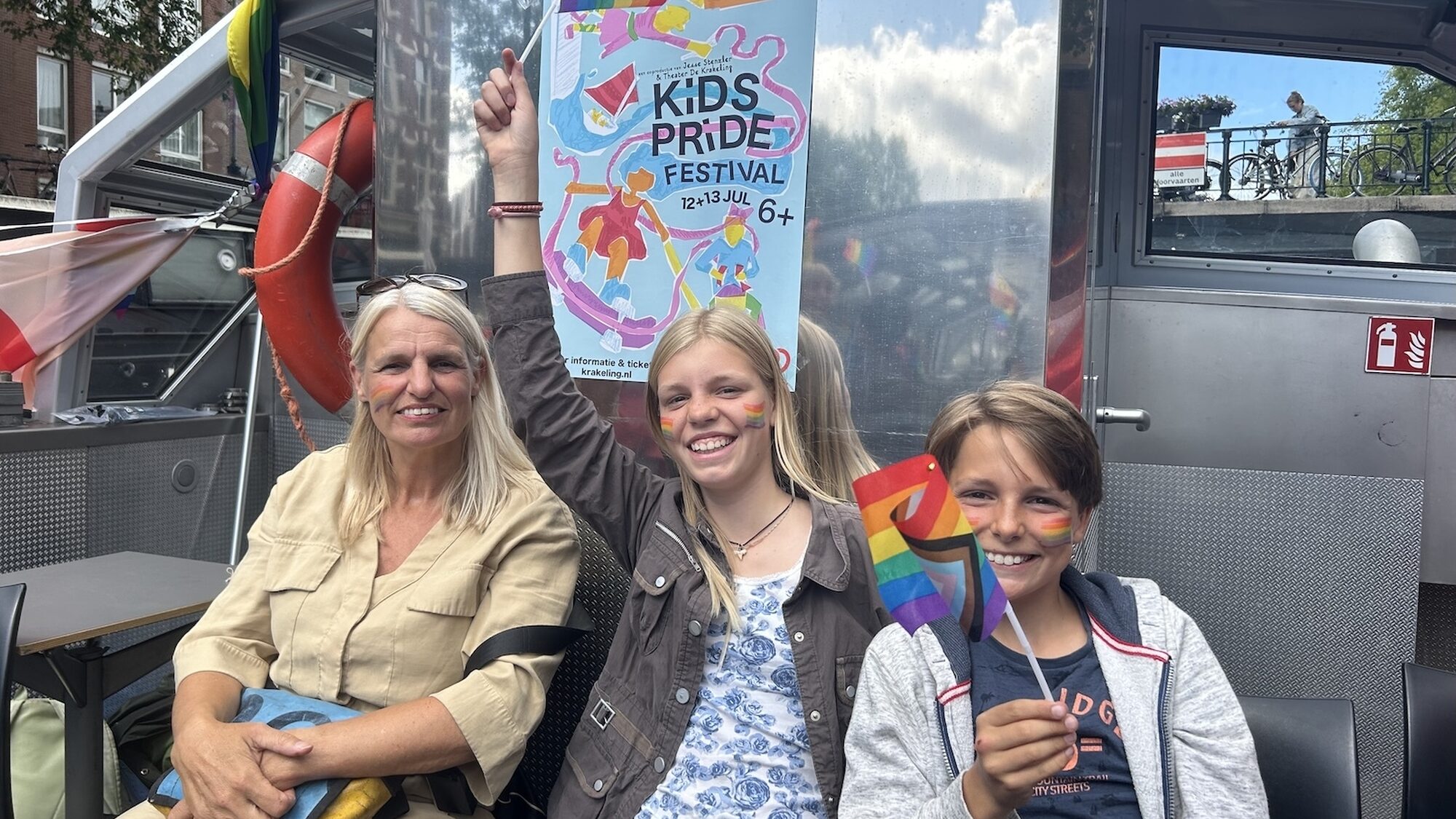 Een moeder en twee kinderen aan boord van de boot. De kinderen zwaaien met regenboogvlaggetjes.