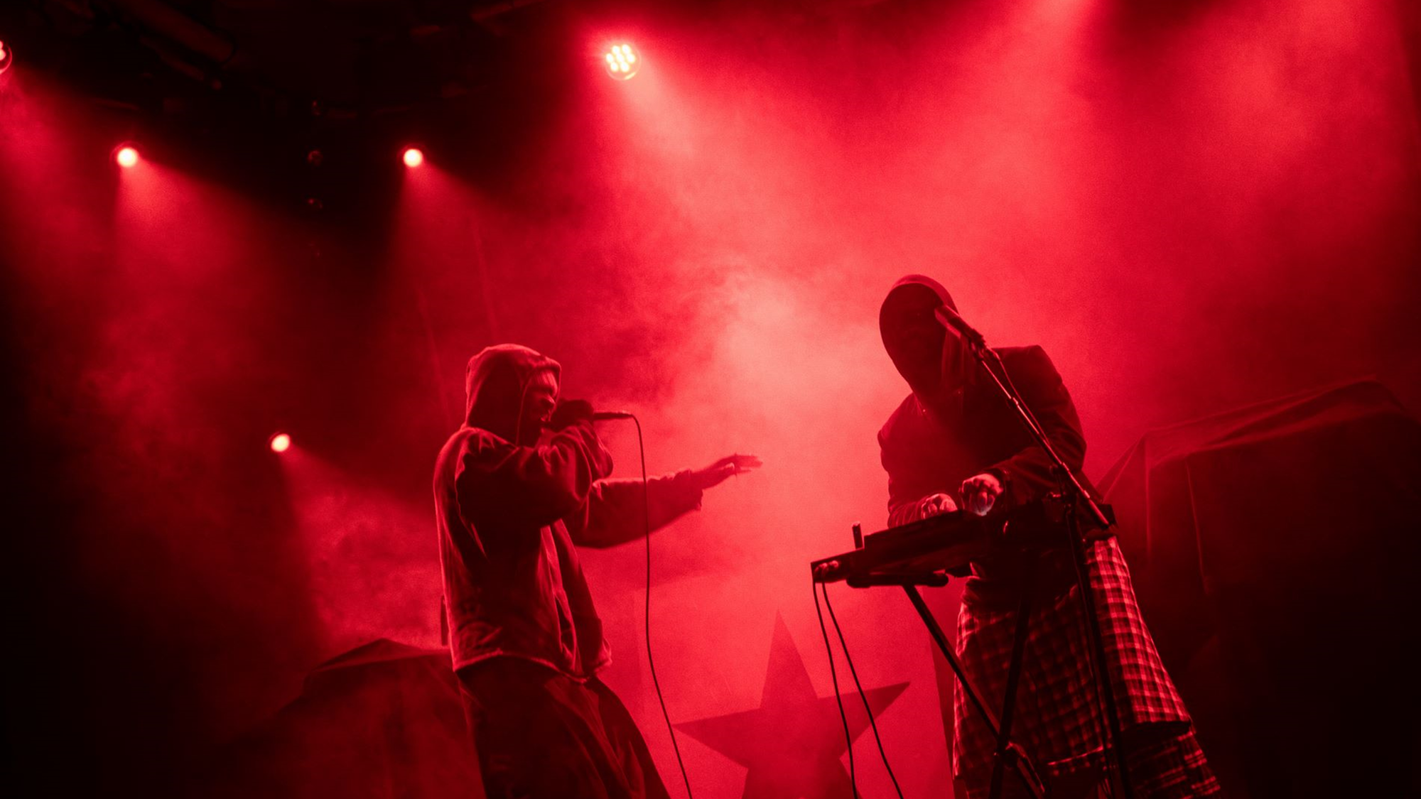 Artiest Isaï staat op het podium op te treden, naast hem staat een man. Het decor bestaat uit rode lampen, rook en een zwarte ster op de achtergrond.
