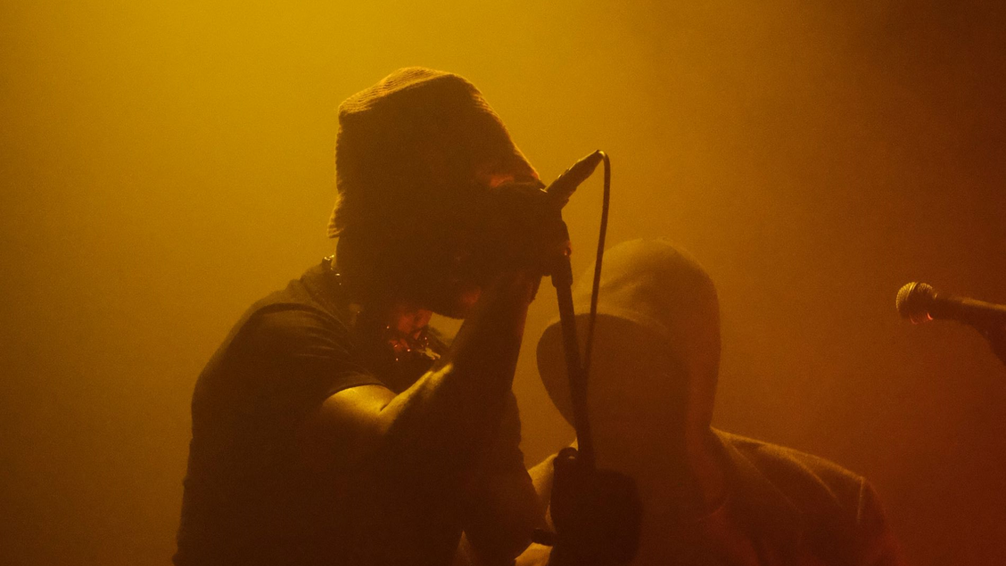 Artiest Isaï staat op het podium op te treden, achter hem staat een man. Het decor bestaat uit gele lampen en veel rook. Beide gezichten van de mannen zijn niet zichtbaar door hoofddeksels.