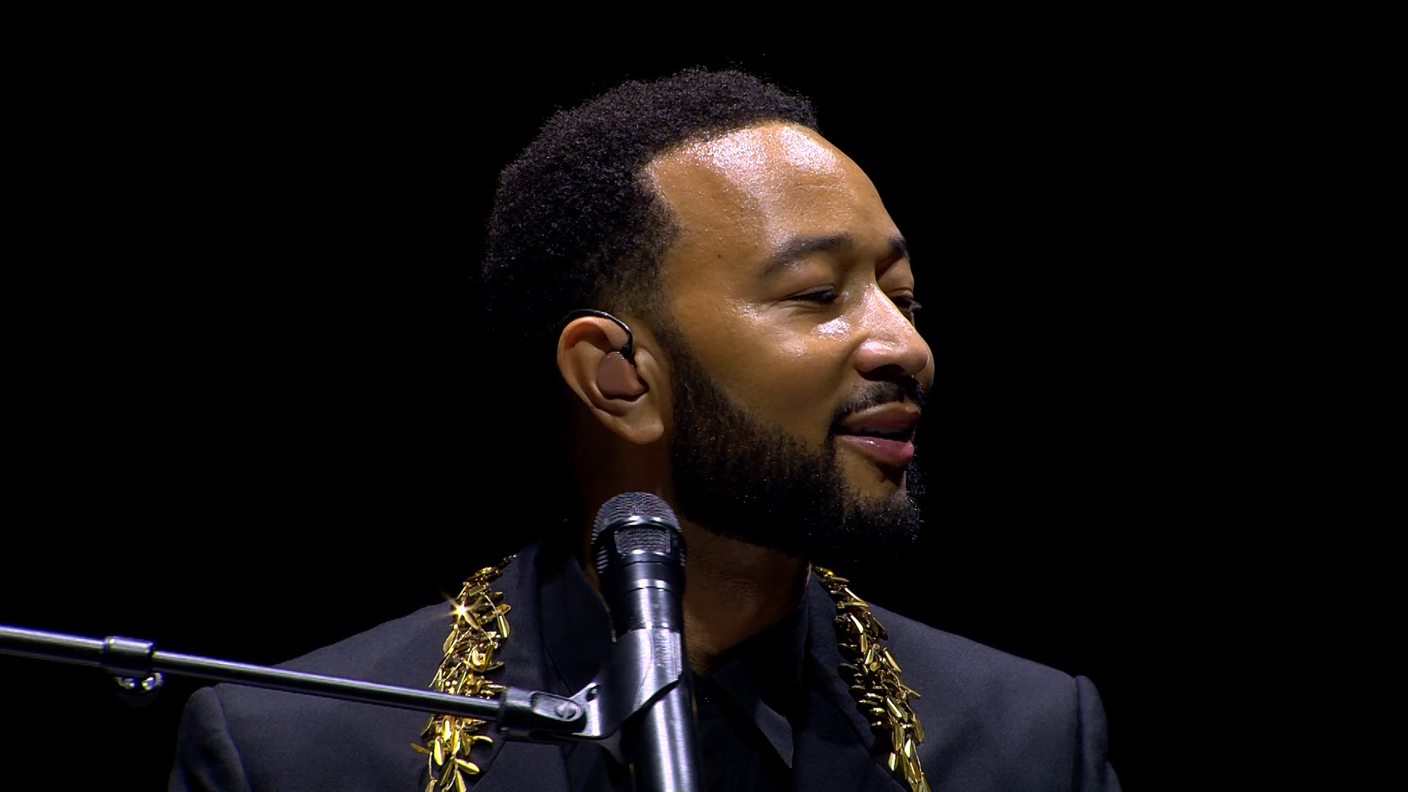 Zanger John Legend zit in een zwart pak met een gouden ketting achter zijn vleugel