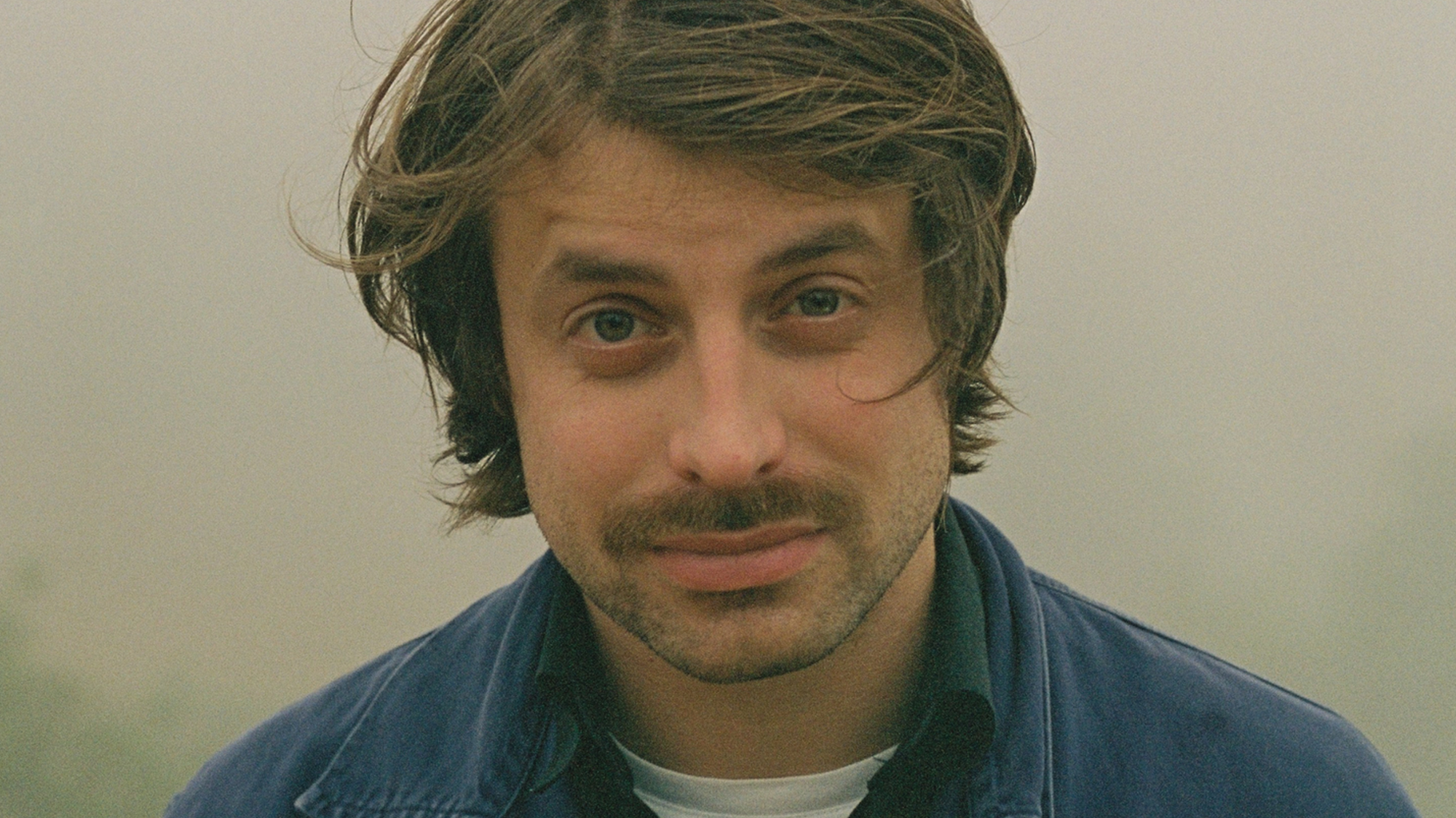 Een portretfoto van Joost Oomen. Een jongeman met wilderig haar, buiten gefotografeerd. Hij heeft een snor en draagt een blauwe jas.