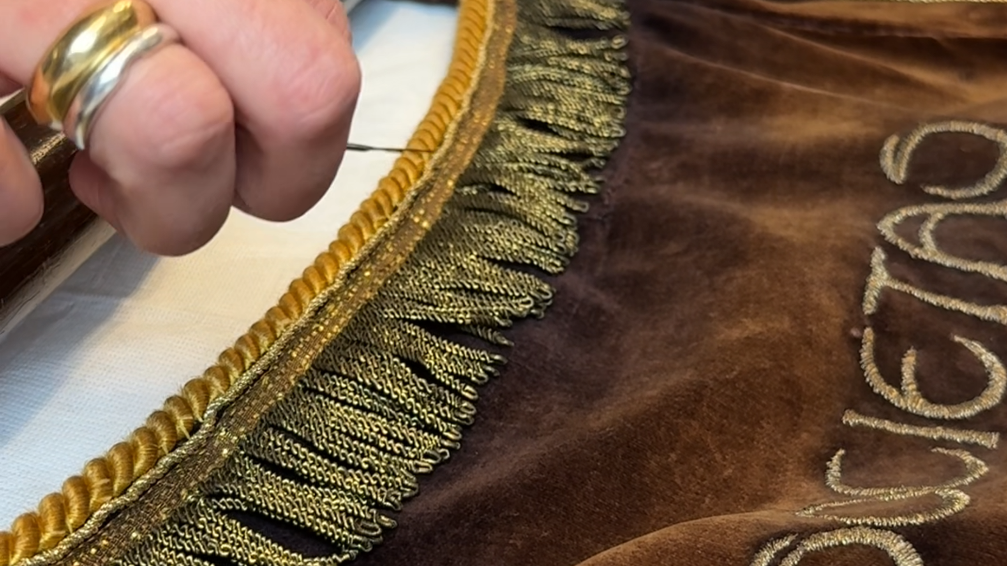 Een close-up van het vaandel. Het is bruine fluweel stof. Je ziet Lorena's hand. Er zitten kleine kwastjes aan het vaandel, die zij aan de kant schuift met een metalen instrument.