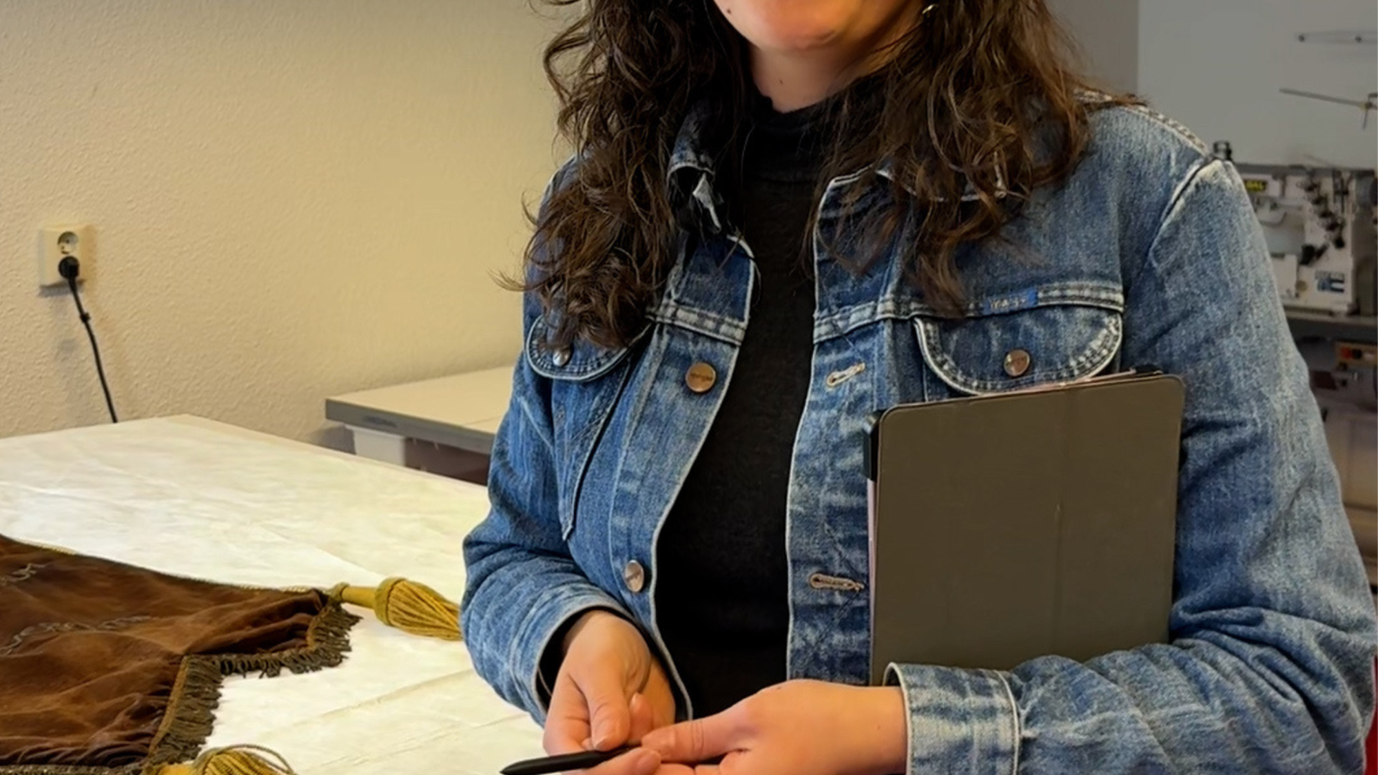 Lorena in haar atelier. Ze heeft een spijkerpak aan en los bruin haar. Ze lacht naar de camera. Ze heeft een tablet en pen in haar hand.