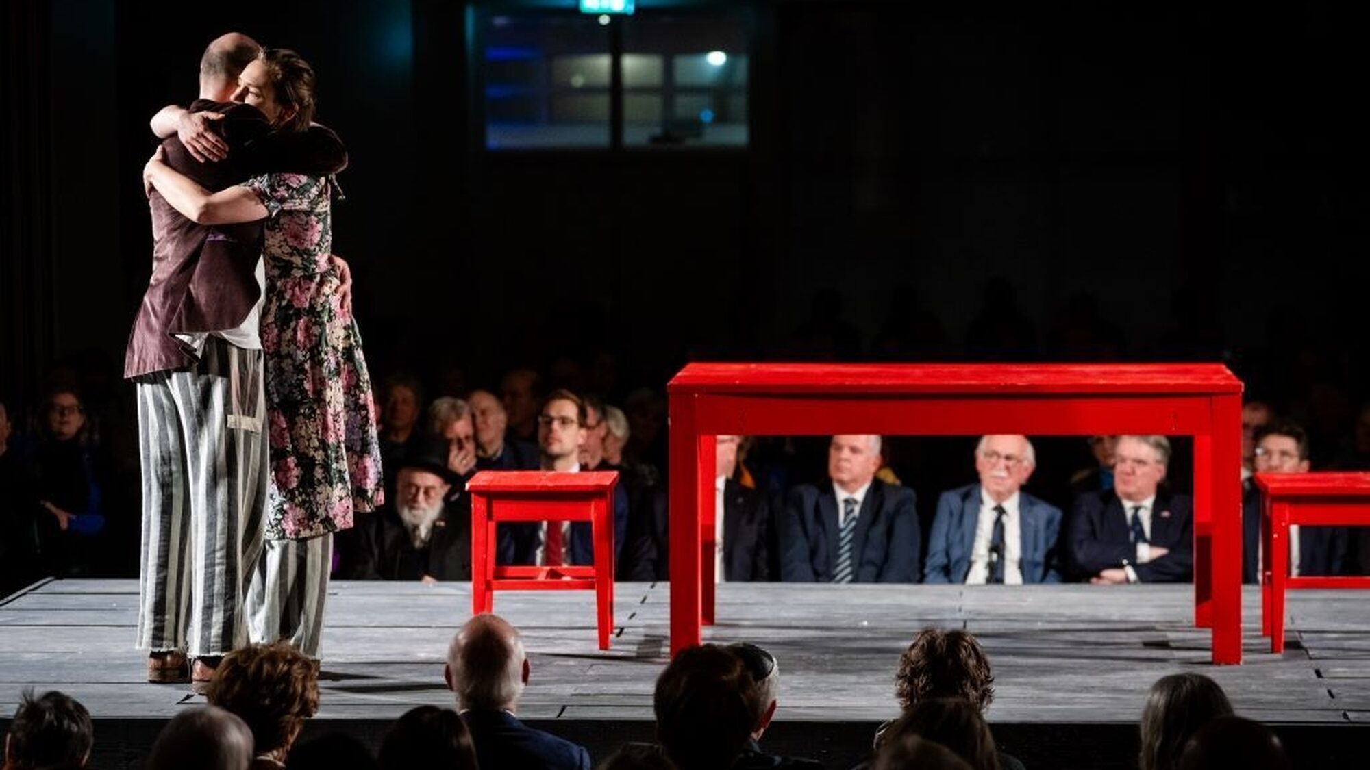 Op de foto zijn twee knuffelende personen op een podium te zien. Een persoon, de vrouw, heeft een bloemetjesjurk aan tot over haar knieën. Daaronder draait ze een loszittende grijs-gestreepte broek. De man draagt een bruin colbert. Ook hij draait dezelfde soort grijs-gestreepte broek. Naast hen op het podium staan een rode tafel en stoel.
