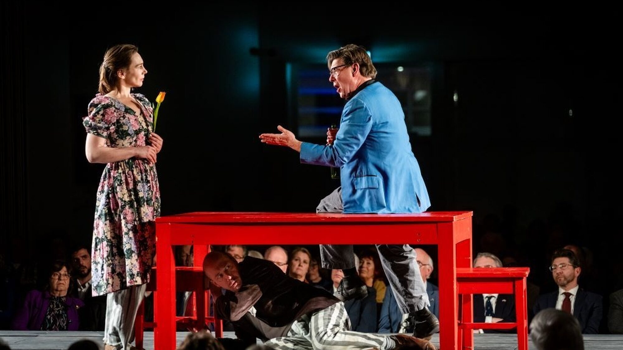 Hier zie je op een podium drie mensen staan rondom, op of onder een rode tafel en twee rode stoelen. Links van de tafel staat een vrouw met een bloemetjesjurk aan en daaronder een gestreepte broek. Ze heeft een bloem in haar handen. Op de tafel zit een man met een blauw colbert en daaronder een gestreepte broek. Hij heeft zijn mond open want hij zingt en hij wijst met een hand naar de vrouw. Onder te tafel ligt een man op zijn zij. Hij draagt een bruin colbert en dezelfde gestreepte broek.