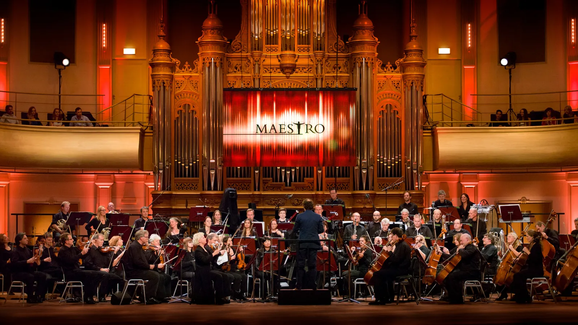 Groot klassiek orkest zit op het podium te spelen met maestro groot op het scherm achter het orkest