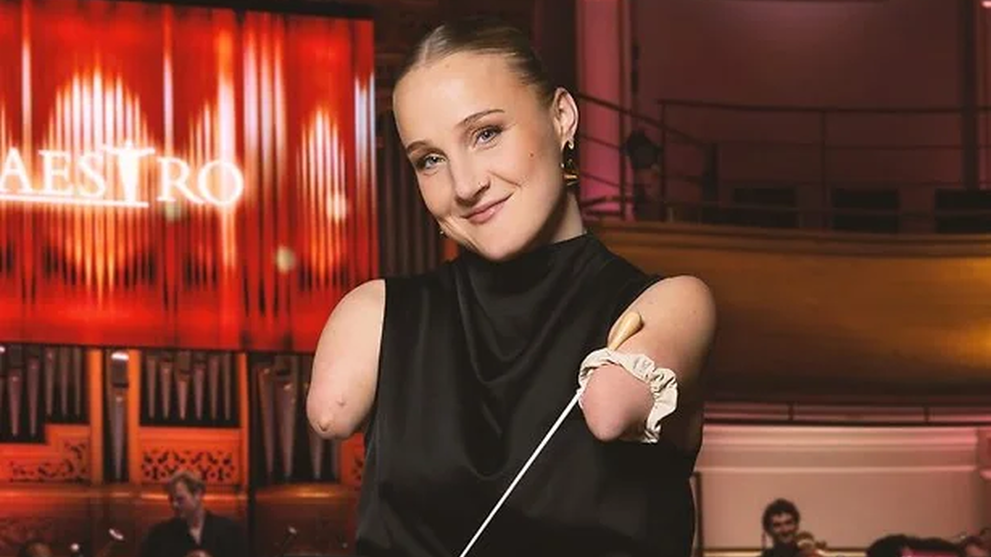 Jonge mevrouw staat met een stokje klaar om te dirigeren voor het orkest van maestro