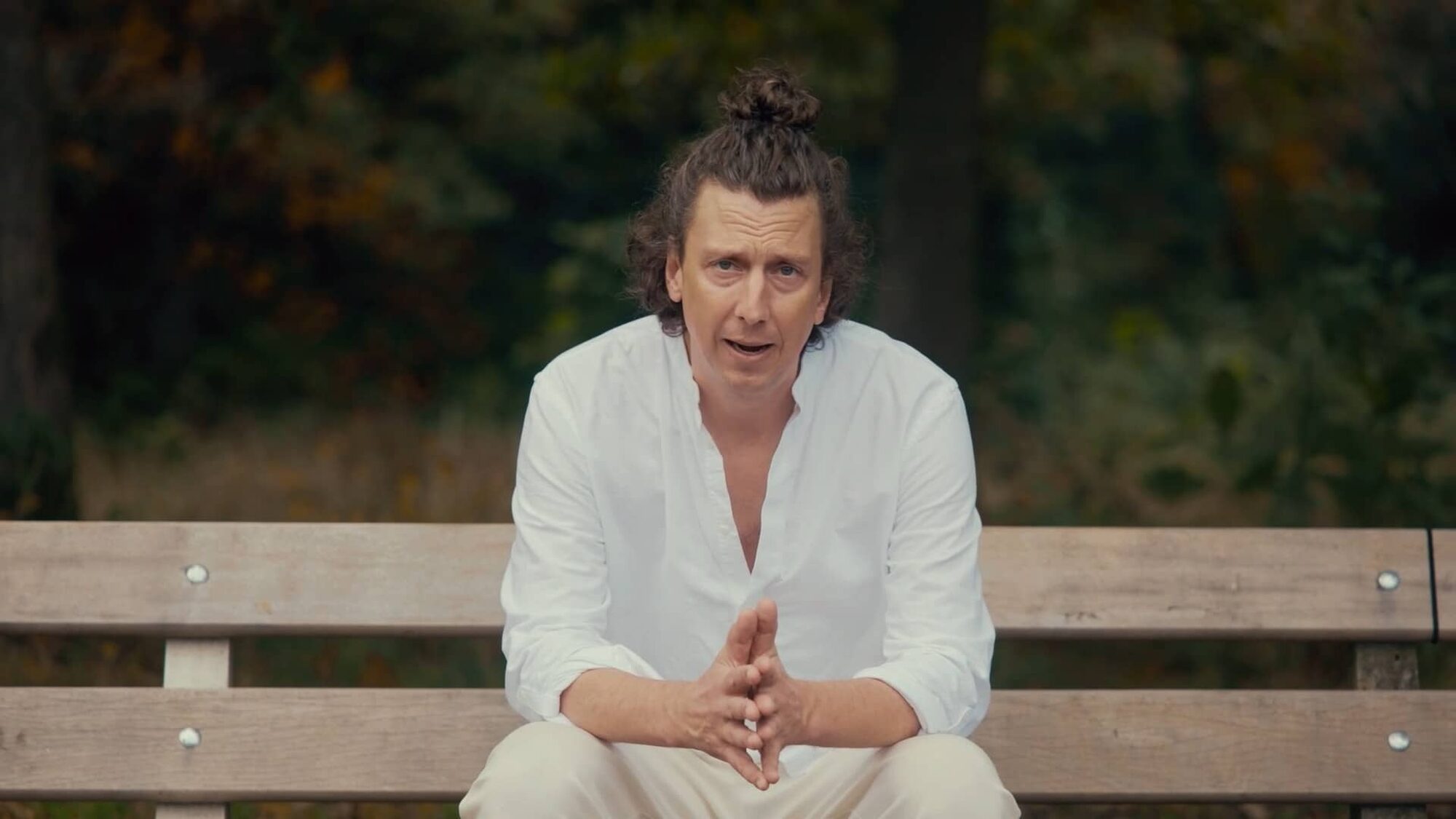 Man met een knot en een wit overhemd zit op een bankje in een het park kijkend naar de camera