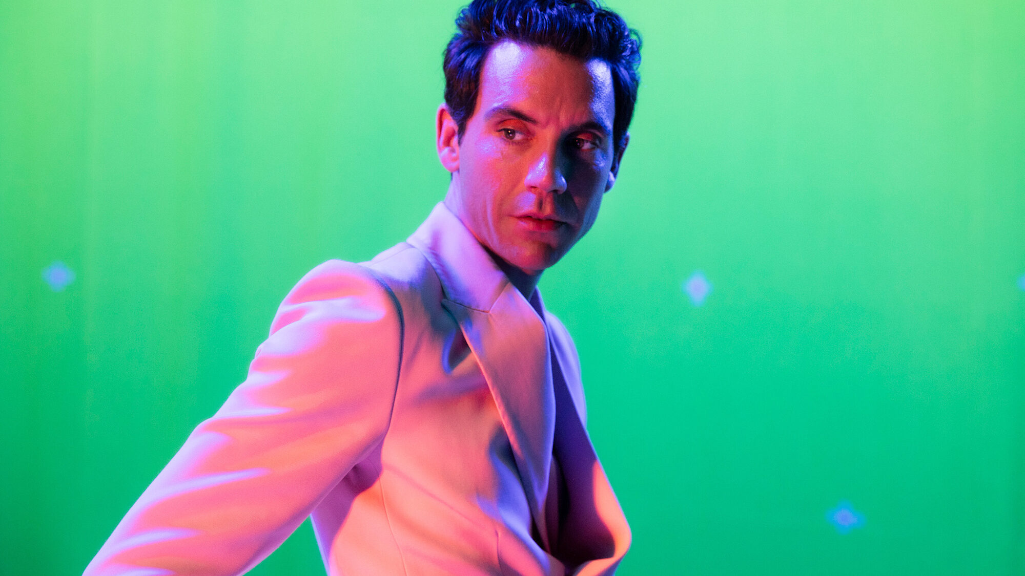 Foto van zanger Mika, in een roze pak voor een groene achtergrond.