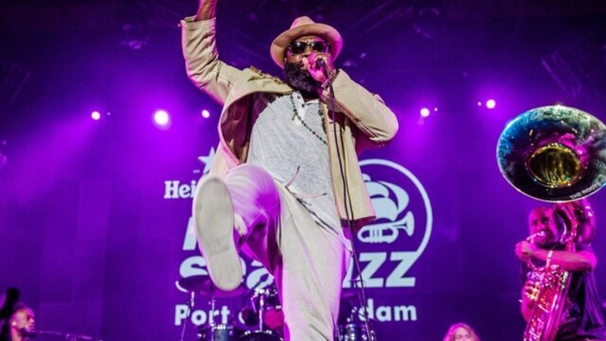 Een zanger danst en zingt op het podium van North Sea Jazz. Achter hem staan muzikanten en het podium is paars verlicht.