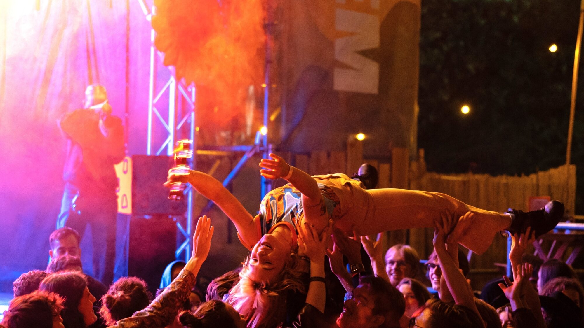 Nelleke crowdsurft over het publiek, ze heeft haar handen omhoog en wordt gedragen door tientallen mensen. De lichten op het podium schijnen deels in de camera waardoor er in de linkerhoek een geel lichteffect ontstaat.