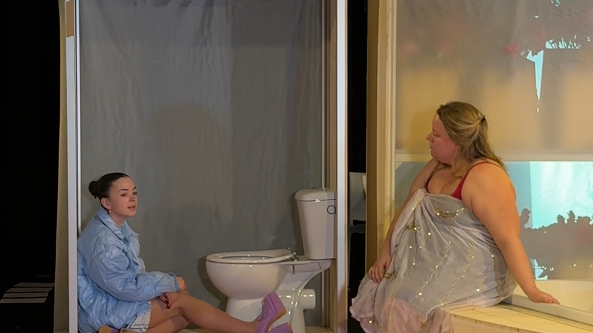 De twee jongere actrices uit het stuk zitten samen in het decor. De ene zit naast een wc en de ander op de badrand.