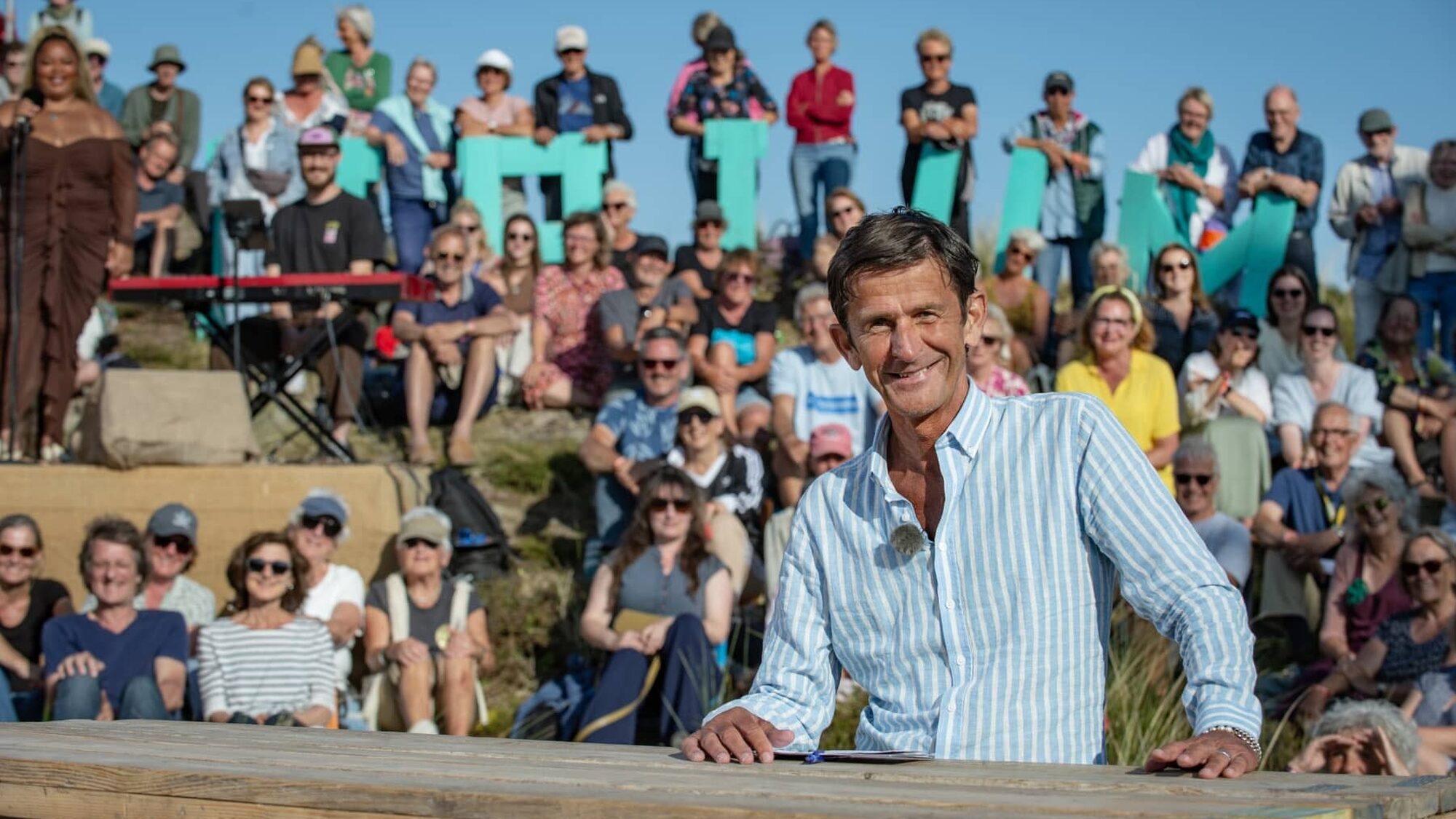Cornald Maas staat glimlachend in een overhemd achter zijn tafel op de set van Opium op Oerol op Terschelling met heel veel mensen achter hem