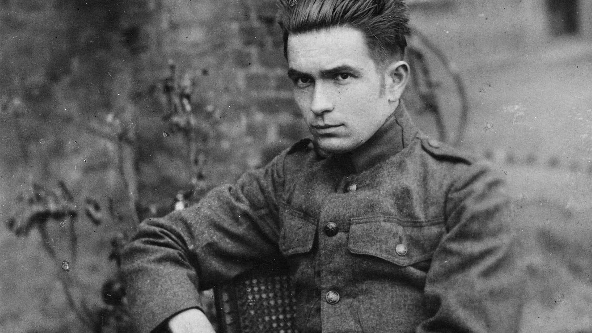 Zwart wit foto met witte man met omhoog gekamd haar zit in militair uniform op een stoel buiten op het gras