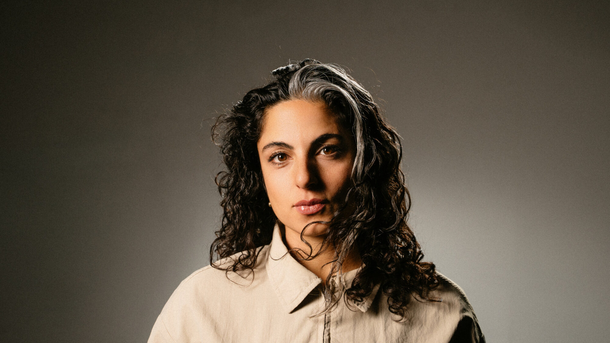 Een foto van Yeliz, de actrice over wie dit stuk gaat. Ze kijkt te camera in en staat voor een bruine achtergrond. Haar zwarte krullen hangen los langs haar hoofd en ze draagt een lichtbruine blouse.