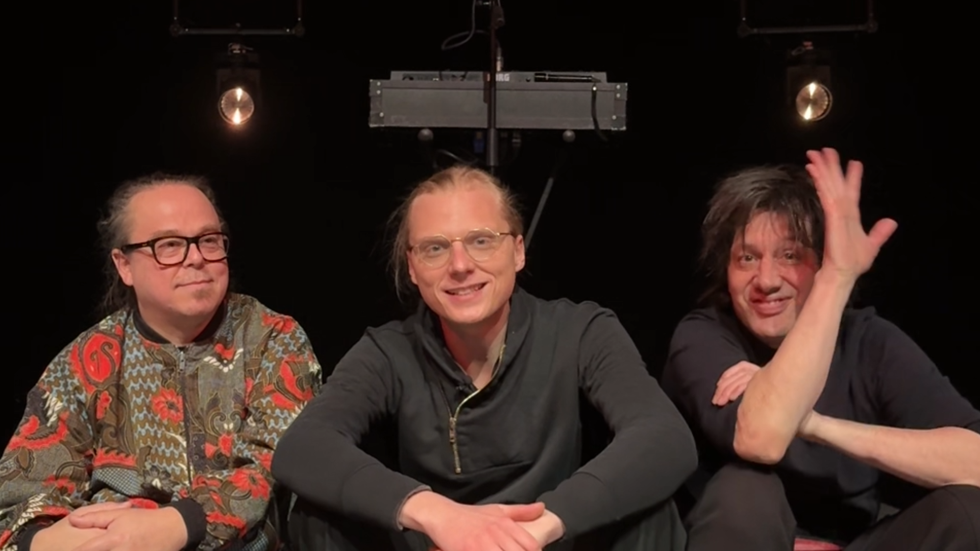 De drie leden van de band Prins S en de Geit zitten op een klein podiumpje en kijken naar de camera. Ze lachen allemaal vriendelijk en de meest rechter zwaait.