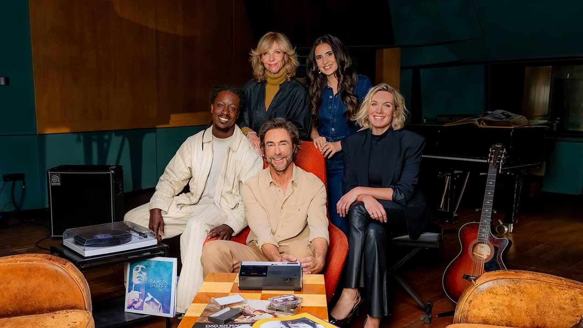 Presentator Chris Zegers zit met de artiesten Claudia de Breij, Akwasi, Karsu en Marlijn Weerdenburg naast elkaar lachend naar de camera kijkend