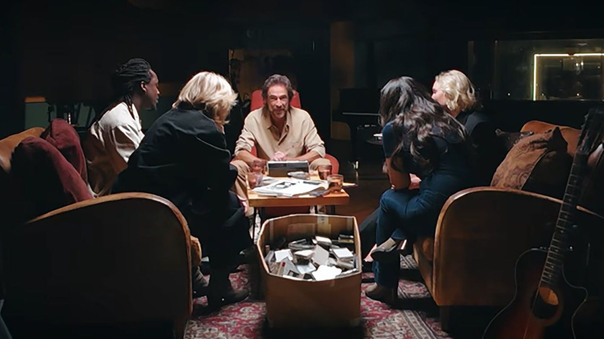 Presentator Chris Zegers zit centraal belicht op een stoel en de vier andere artiesten zitten met hun rug naar de camera aandachtig te luisteren naar een cassetterecorder