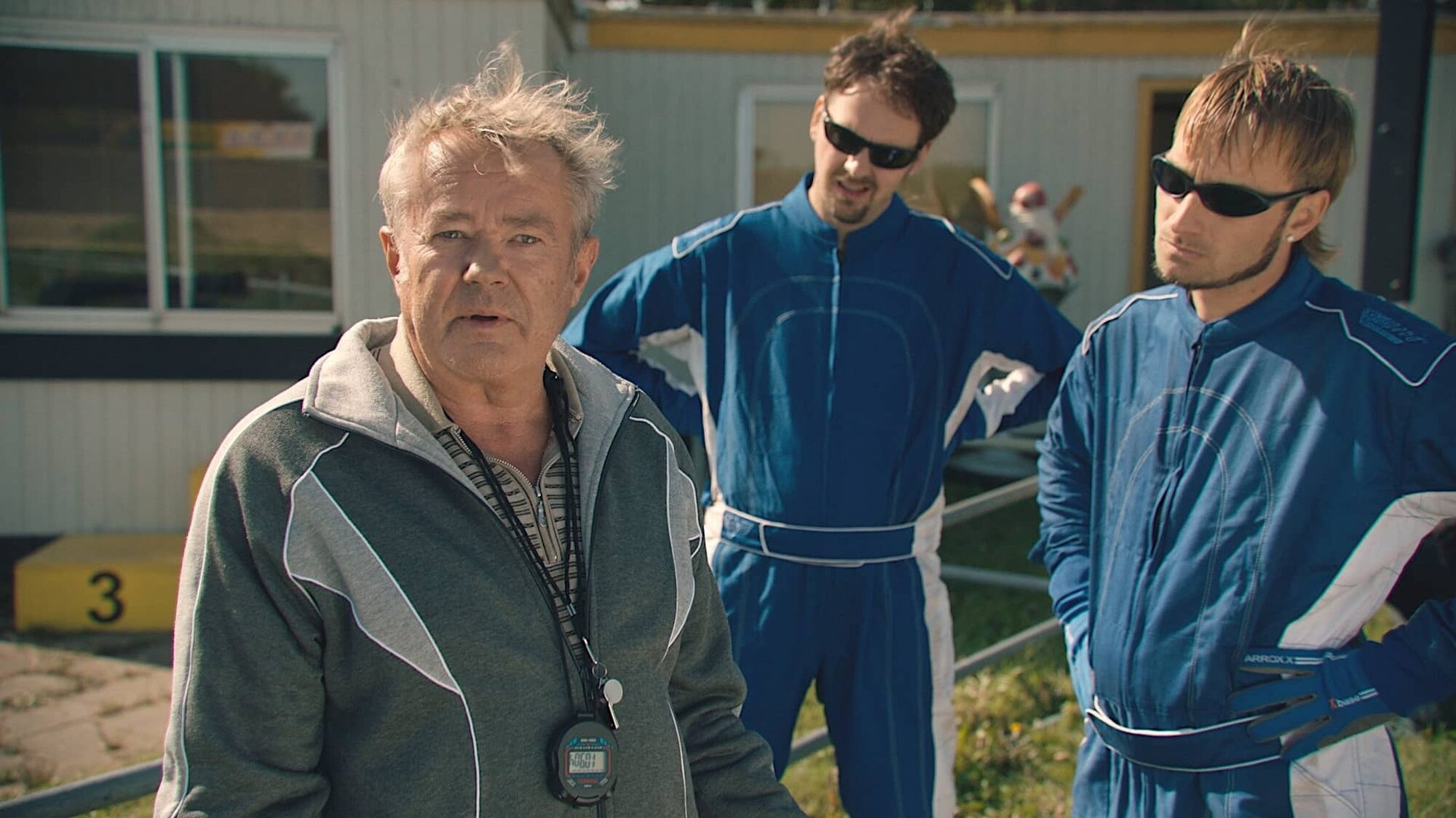 Drie witte mannen spelen hun rol in de serie Schwalbe. Links staat de wat oudere trainer en rechts staan de twee stuurlopers in blauwe pakken met zwarte zonnebrillen.