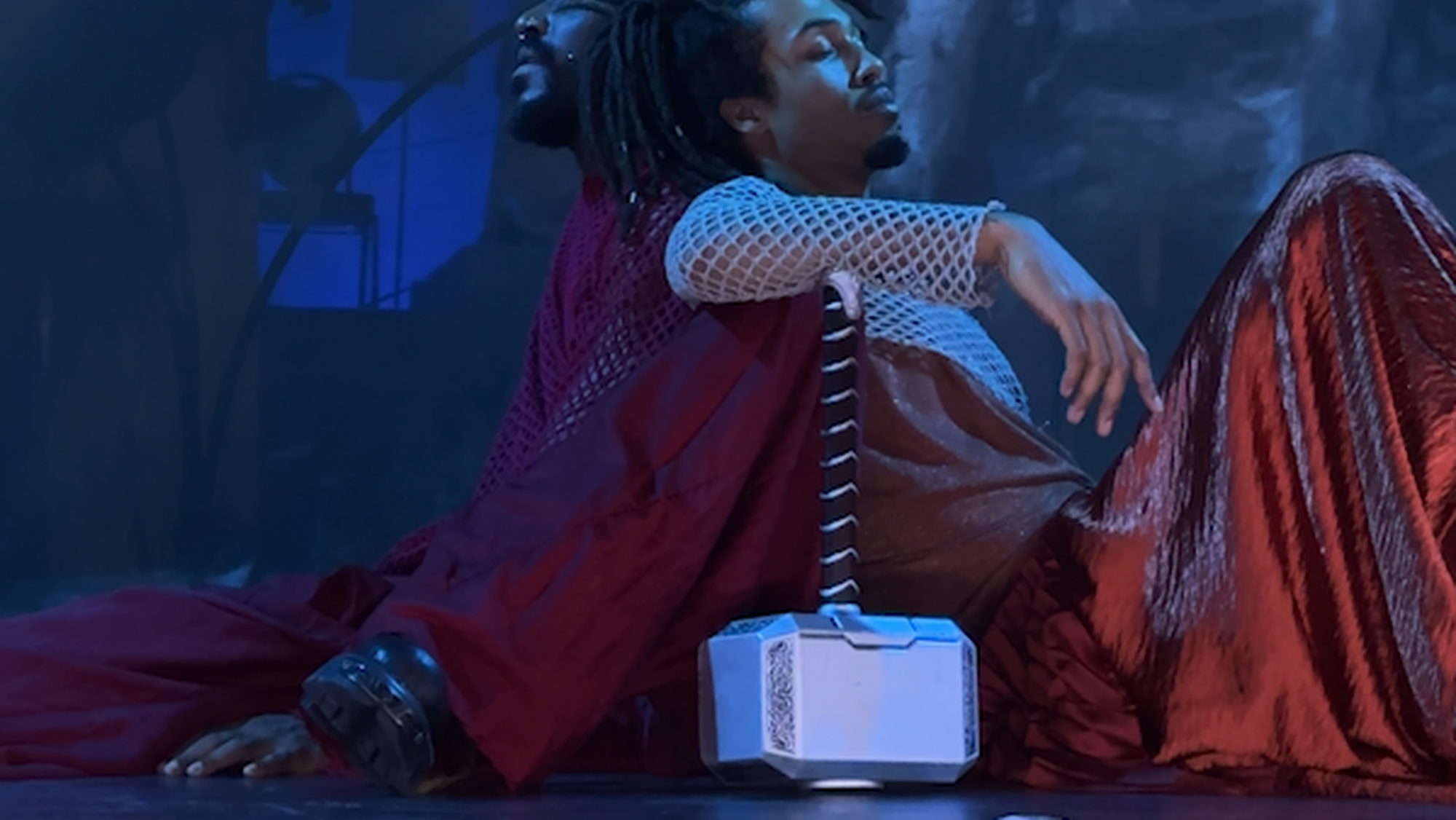 je ziet 2 acteurs van kleur met de grijze ijzeren hamer van Thor, de Noorse god. zij vertolken de rollen van Thor en Loki in oranje en rode gewaden.