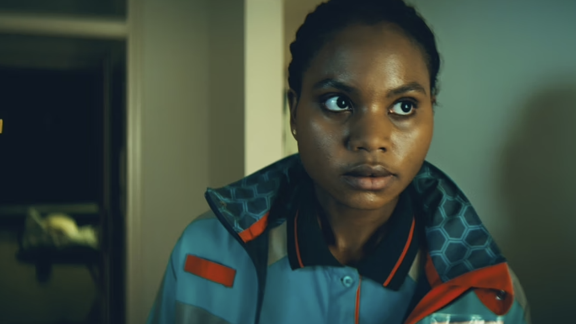 Alima staat in een huis met een ambulance jas aan en kijkt naar iets buiten beeld