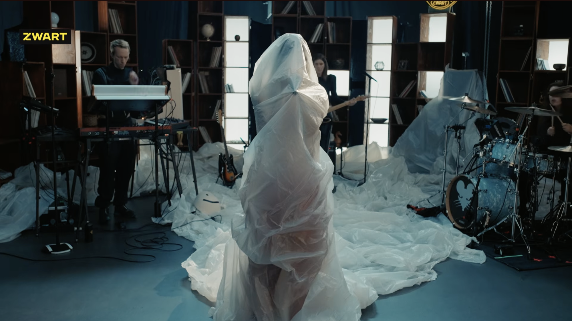 Zangeres MEROL staat in een studio onder een plastic zeil met haar band om haar heen