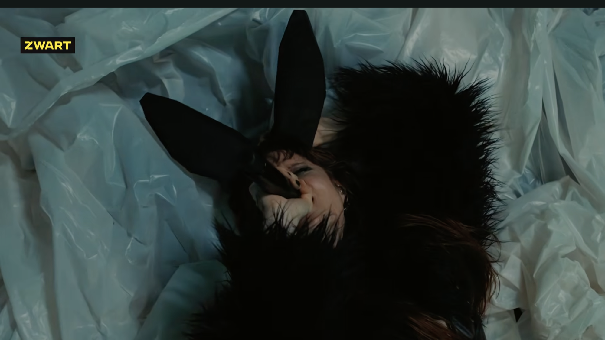 Vrouw met konijnenoren en zwart harige top zingt in een microfoon liggend op de grond