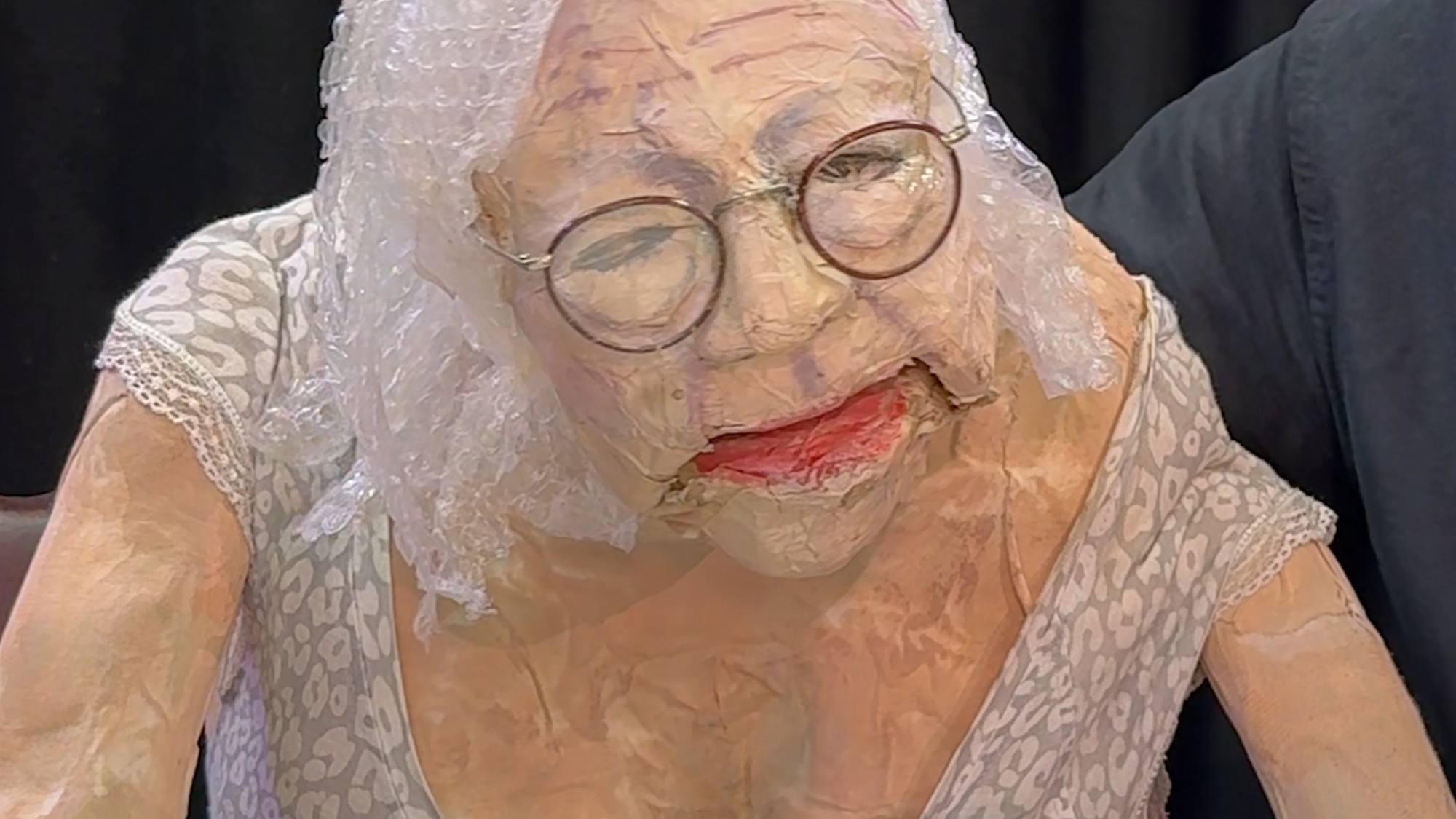 Een foto van de pop. Het is een oude vrouw met een bleke huid, bril en bubbeltjesplastic als haar. Ze heeft een bloemenjurkje aan en haar hand zit aan een theekopje met oranje en bruine strepen. Ze is van een papierachtig materiaal gemaakt.