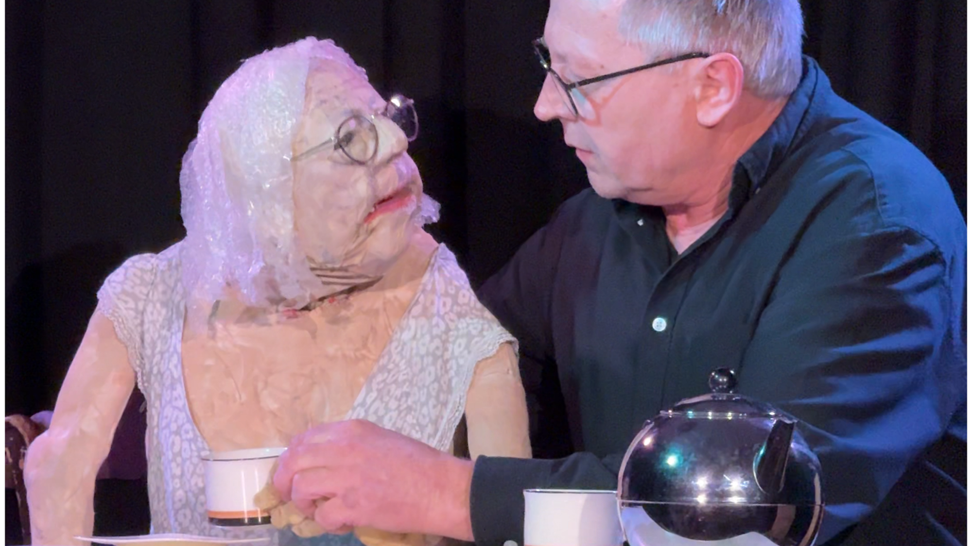 Een foto van Servaes en de pop van zijn moeder achter een tafel. Ze zitten en kijken naar elkaar. Op tafel staat een zilvere, glimmende theepot. Servaes heeft een kopje in zijn hand.