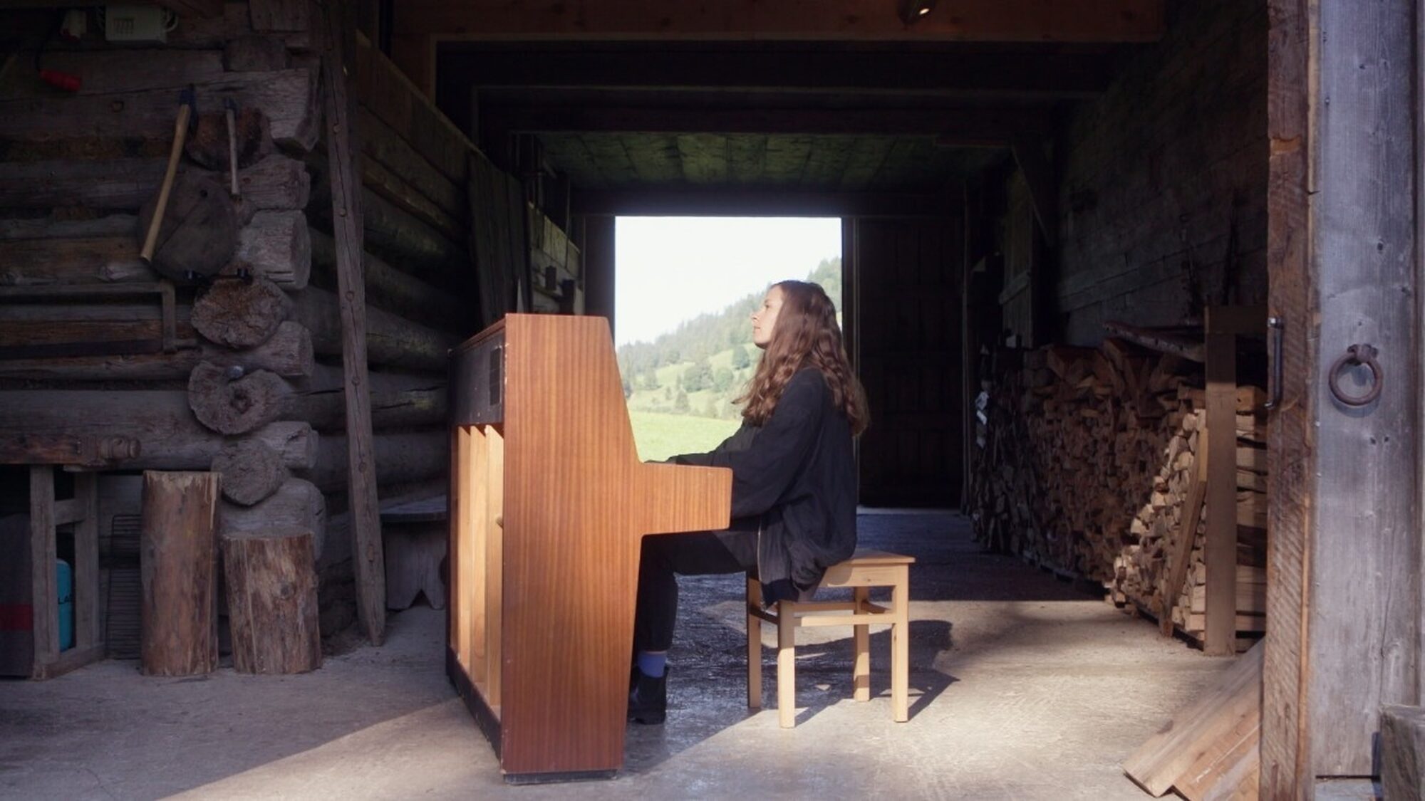 Een vrouw speelt piano in een houten schuur. Achter haar zie je een berg.