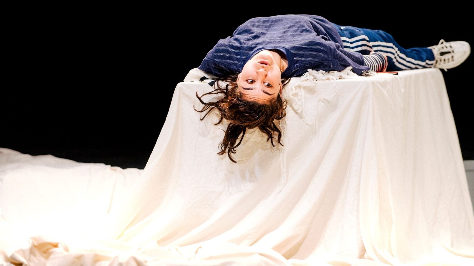 Actrice Soumaya ligt in blauwe kleding met haar rug op een tafel met een groot wit doek. Haar hoofd hangt over de tafel heen en ze kijkt naar rechts.