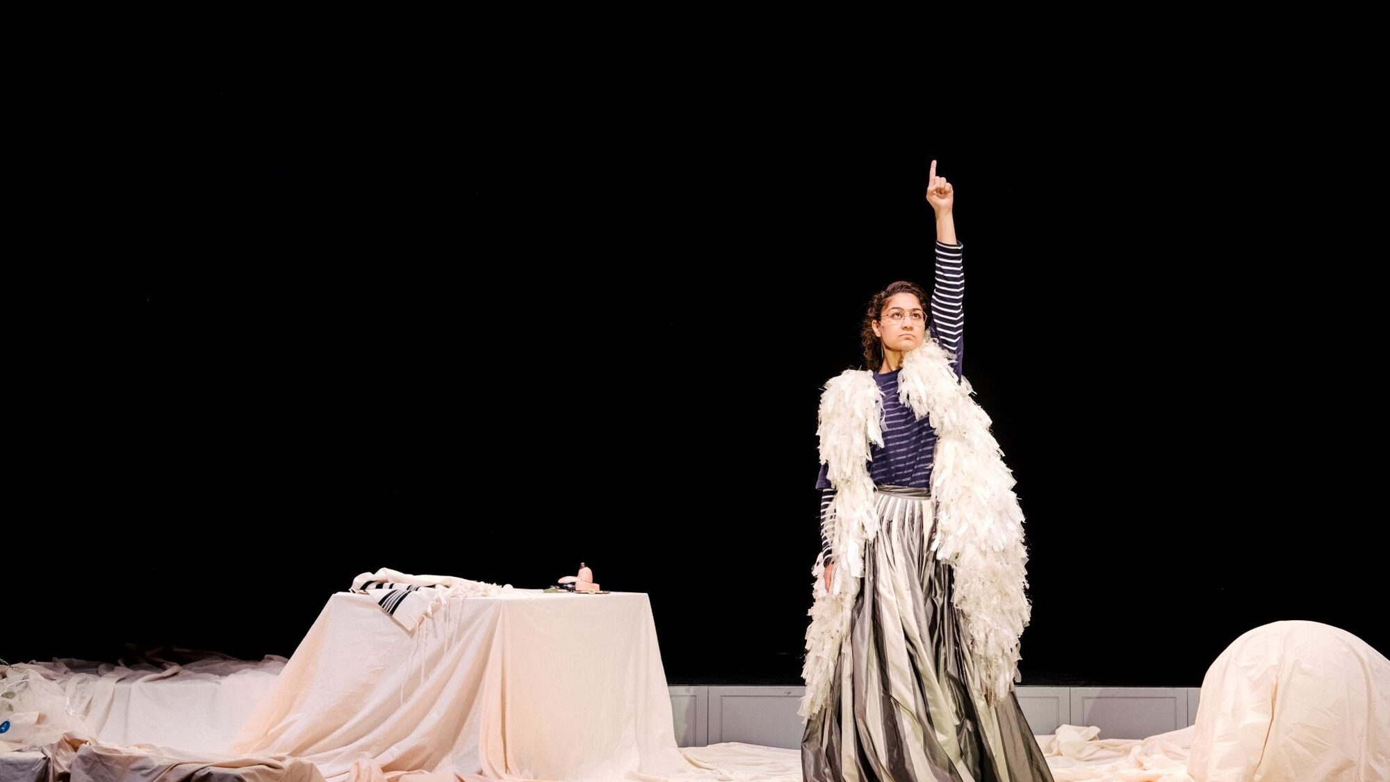 Actrice Soumaya staat op het podium met haar vinger in de lucht. Ze draagt een gestreept shirt, een witte boa en een lange grijze rok. De vloer is bedekt met witte kleden.