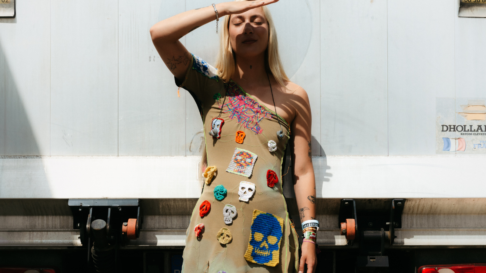 Foto van artiest Stien die voor een vrachtwagen staat en tegen de zon in kijkt. Ze heeft lang blond haar en een jurk vol opgenaaide stukjes stof met doodshoofden erop. De jurk is bruin. Ze houdt haar hand half voor haar gezicht om de zon uit haar ogen te houden en heeft haar ogen dicht.