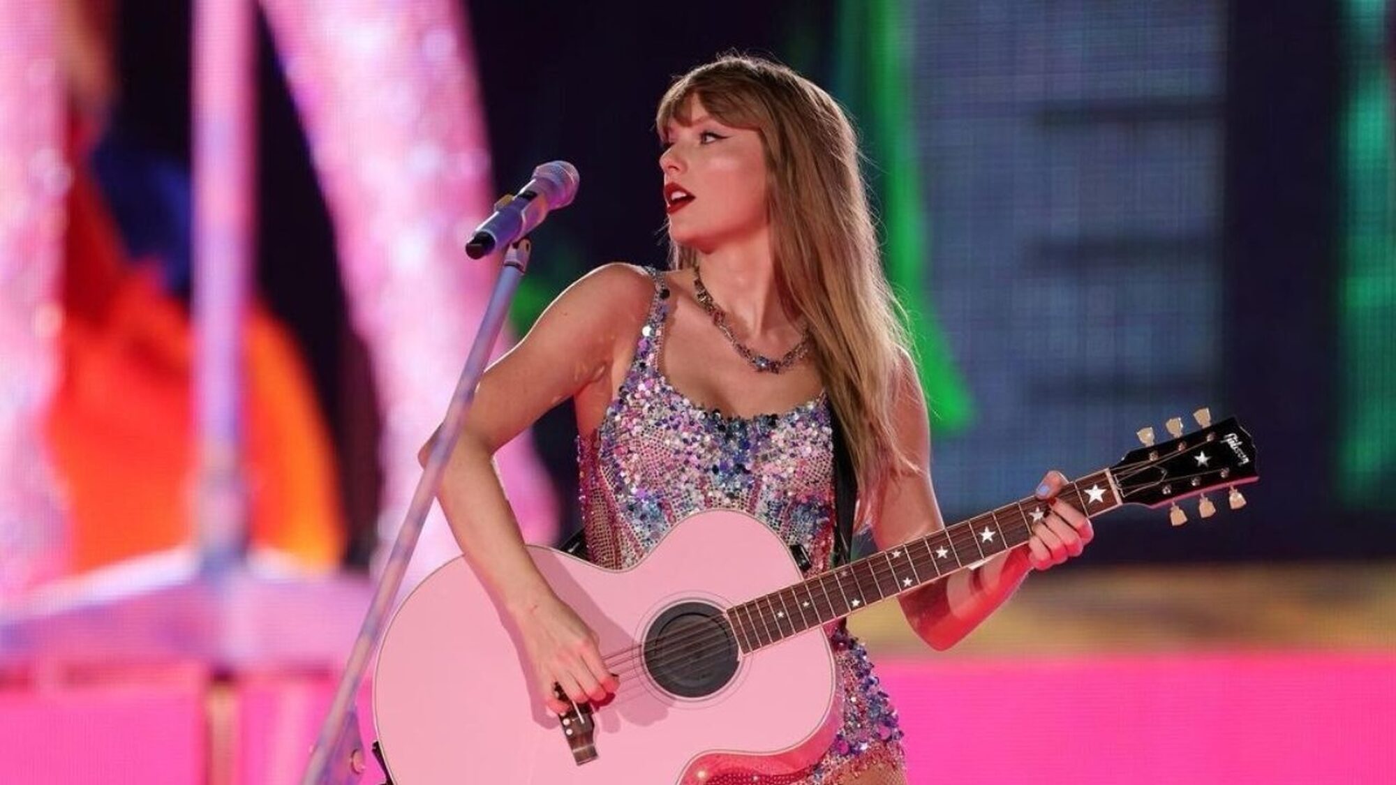 Blonde Amerikaanse zangeres Taylor Swift staat op het podium in een glitterjurkje met een roze gitaar te zingen.