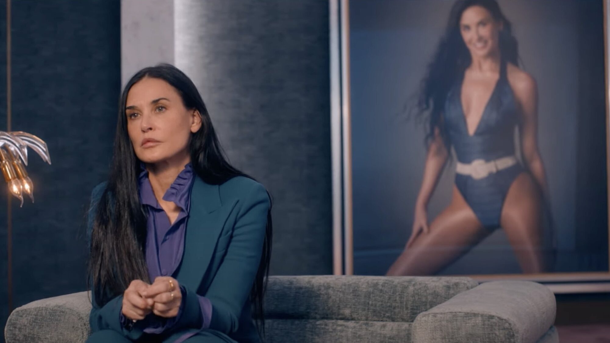Een stil uit de film The Substance. Een treurig kijkende vrouw zit op een bank. ze heeft lang zwart haar en achter haar hangt een geposeerde studio-foto van haar in badpak.