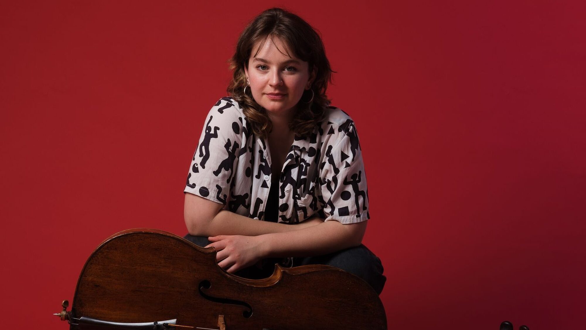 Witte vrouw met bruine haren leunt op een cello kijkend naar de camera met een rode achtergrond