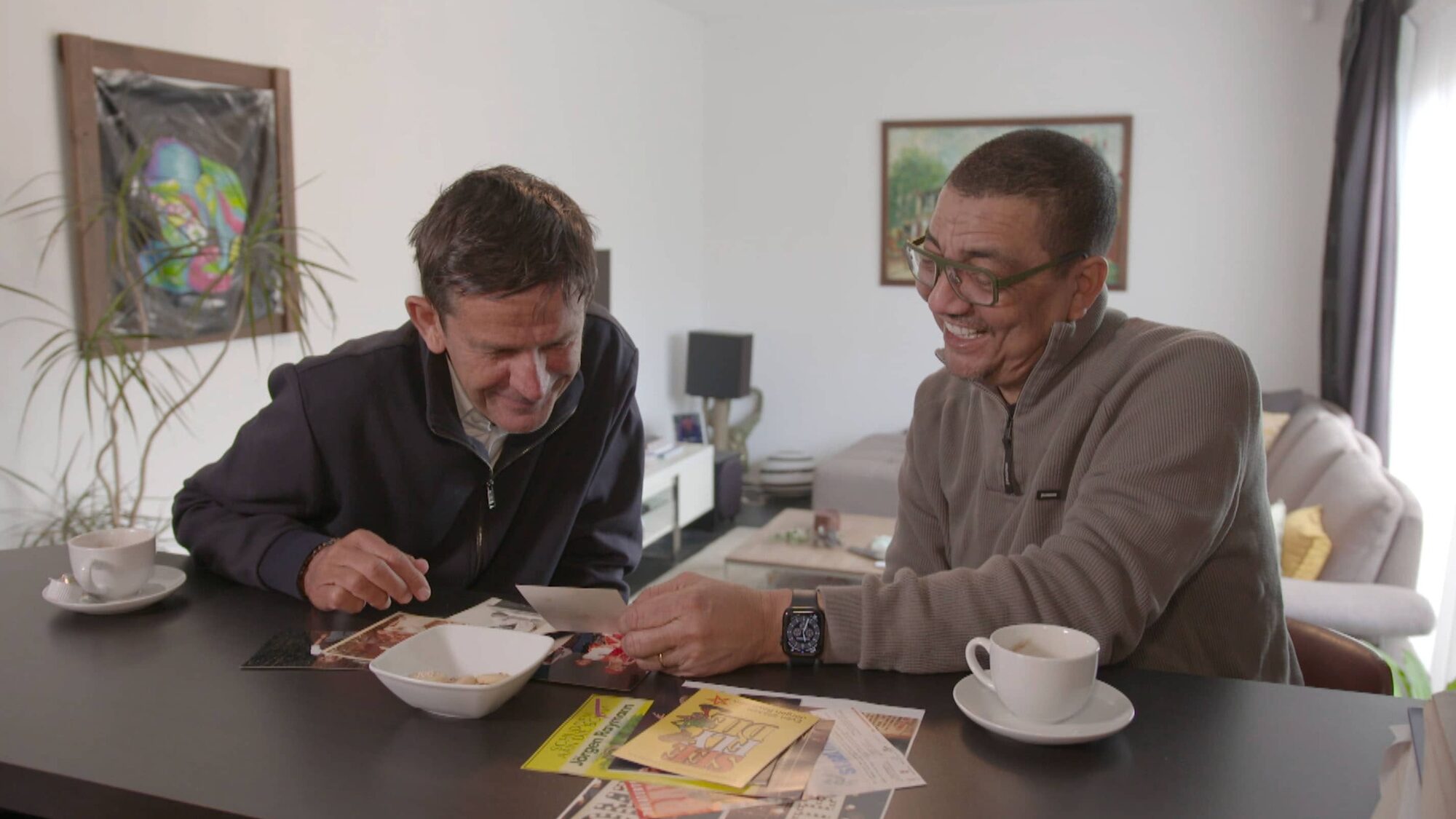 Nederlandse man en Surinaamse man zitten samen aan tafel en lachen om een fotokaartje