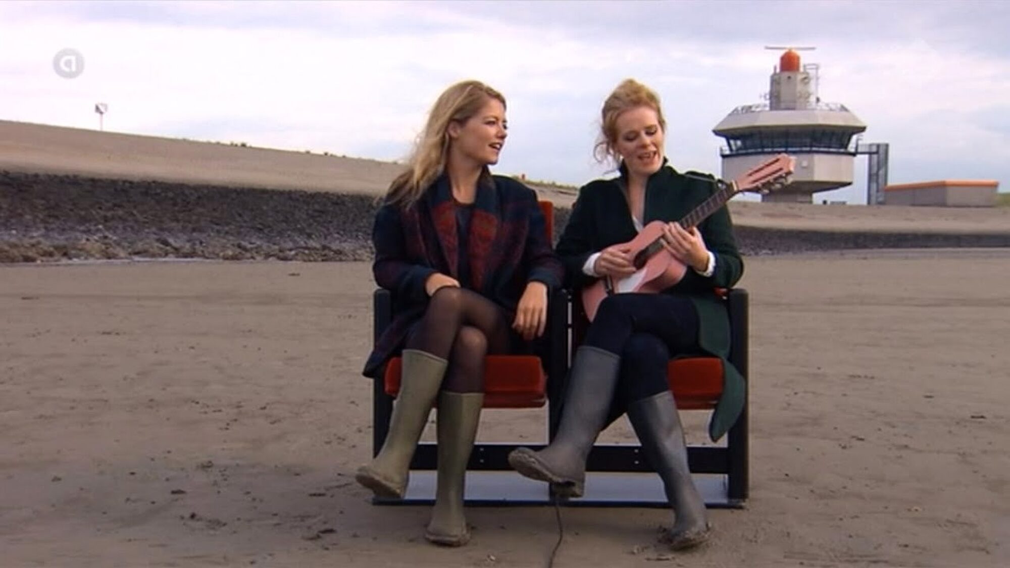 Yentl en de Boer zitten samen op stoeltjes op het strand te zingen met een gitaar