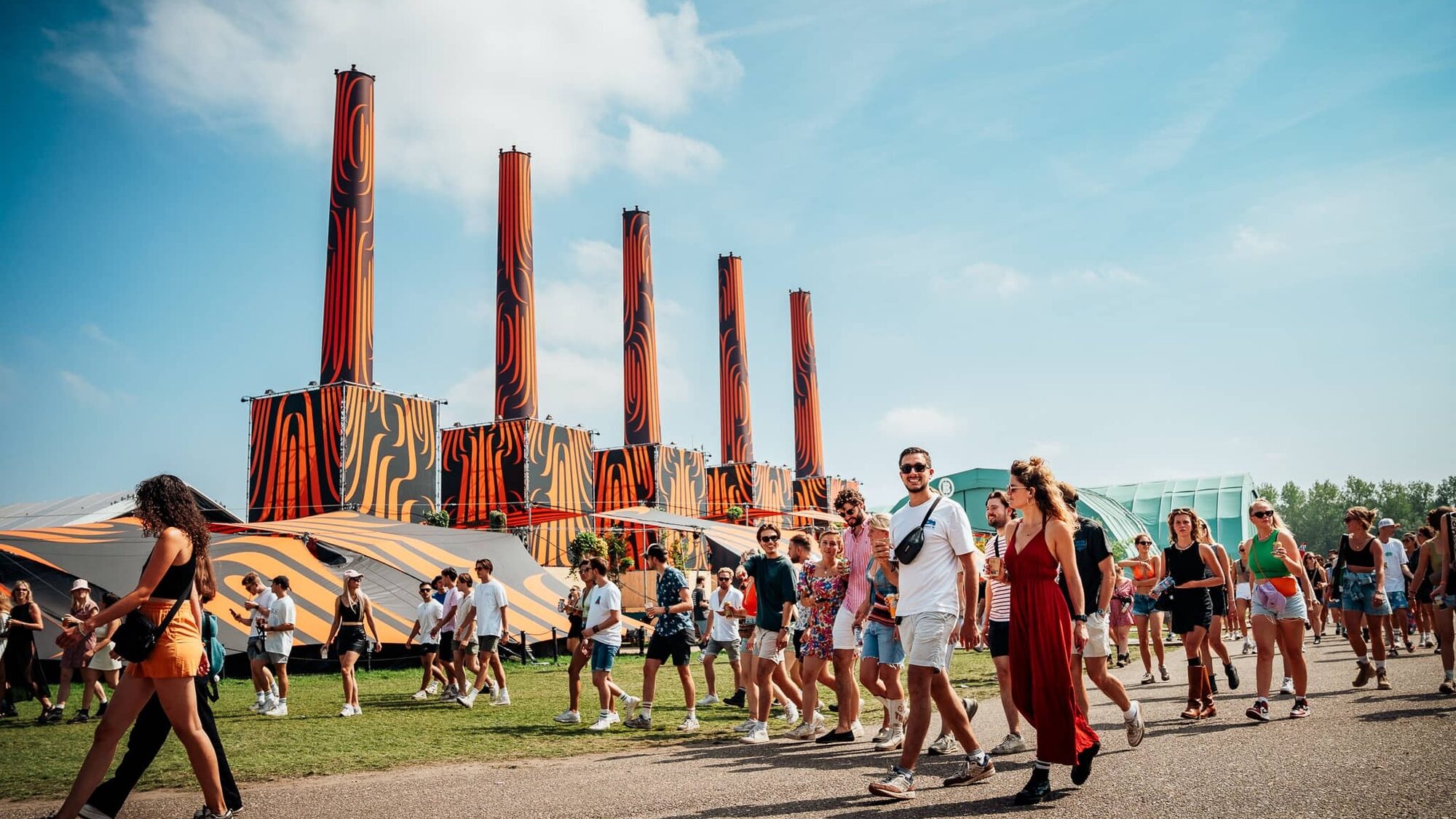 Een foto van het Lowlands festivalterrein. op de voorgrond zie je mensen lopen op asfalt, iedereen draagt kleurrijke en zomerse kleding. Op de achtergrond zie je voor de blauwe lucht een soort tent met grote oranje schoorstenen. Het is een herkenbaar beeld van Lowlands.