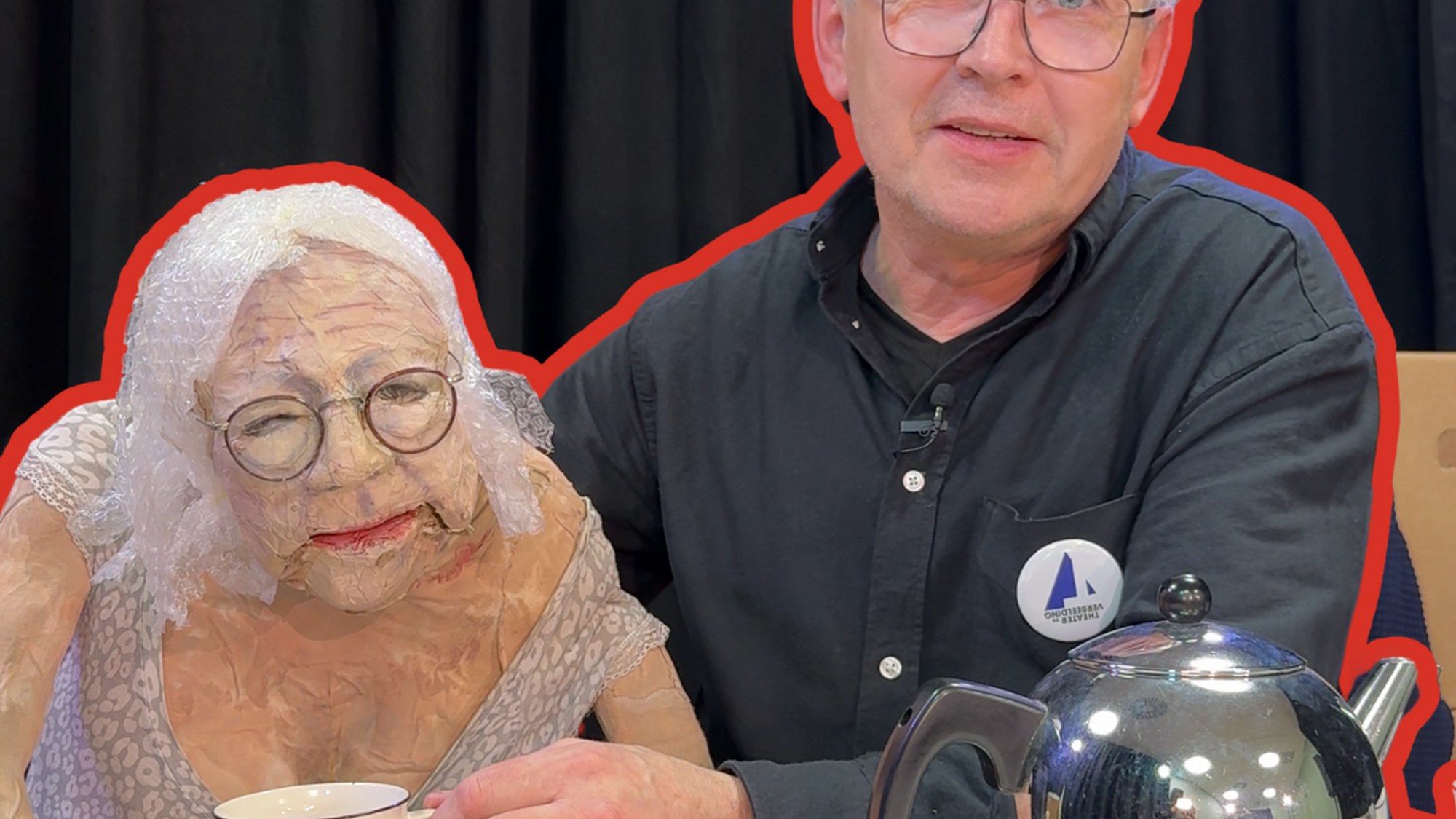 Een foto van Servaes de poppenspeler en de pop die zijn moeder voorstelt. Ze zitten samen achter een witte tafel en er staan een theepot en kopjes. Servaes is een man met een bril en grijs haar en hij draagt een zwart overhemd en kijkt vriendelijk de camera in. De pop naast hem is een oude vrouw, gemaakt van proppen papier en tape, met een bleke huid, bril, bloemenjurk en haar van bubbeltjesplastic.