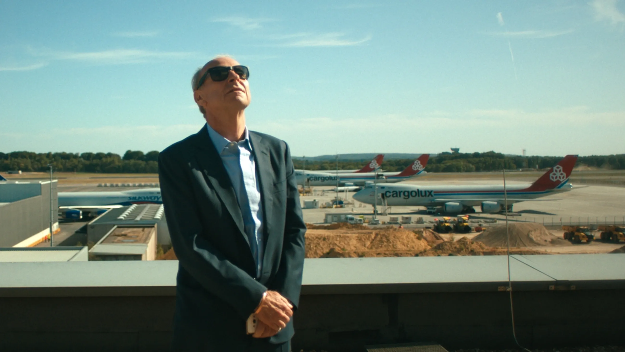 Een stil uit de documentaire. Een oudere man in pak met grote zonnebril op, staat met zijn gezicht in de zon. Op de achtergrond staan meerdere vliegtuigen, het lijkt een klein vliegveld te zijn. Hij heeft zijn handen over elkaar en staat voor een soort balustrade.
