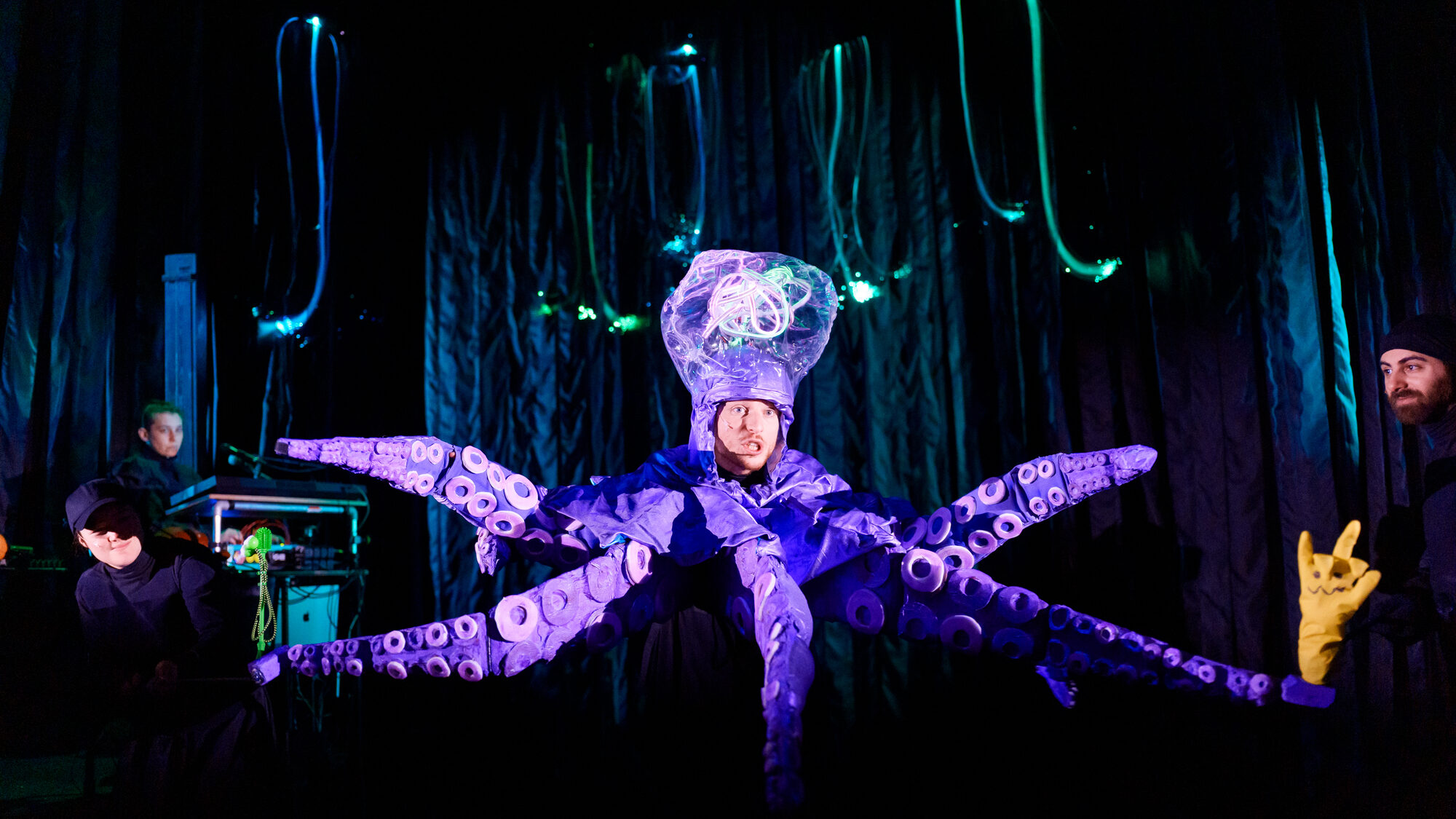 Een foto van de theatervoorstelling DIEP. We zien een man in een octopuspak, voor een donkere achtergrond. Hij lijkt te zingen of praten en heeft fel licht op zijn gezicht. Het octopuspak is paars.
